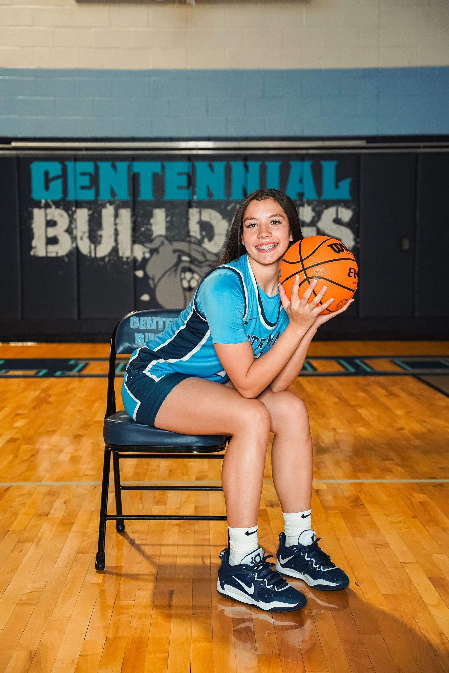 Basketball player wearing turquoise jersey holds orange ball while sitting on gymnasium bench with Centennial Bulldogs signage.