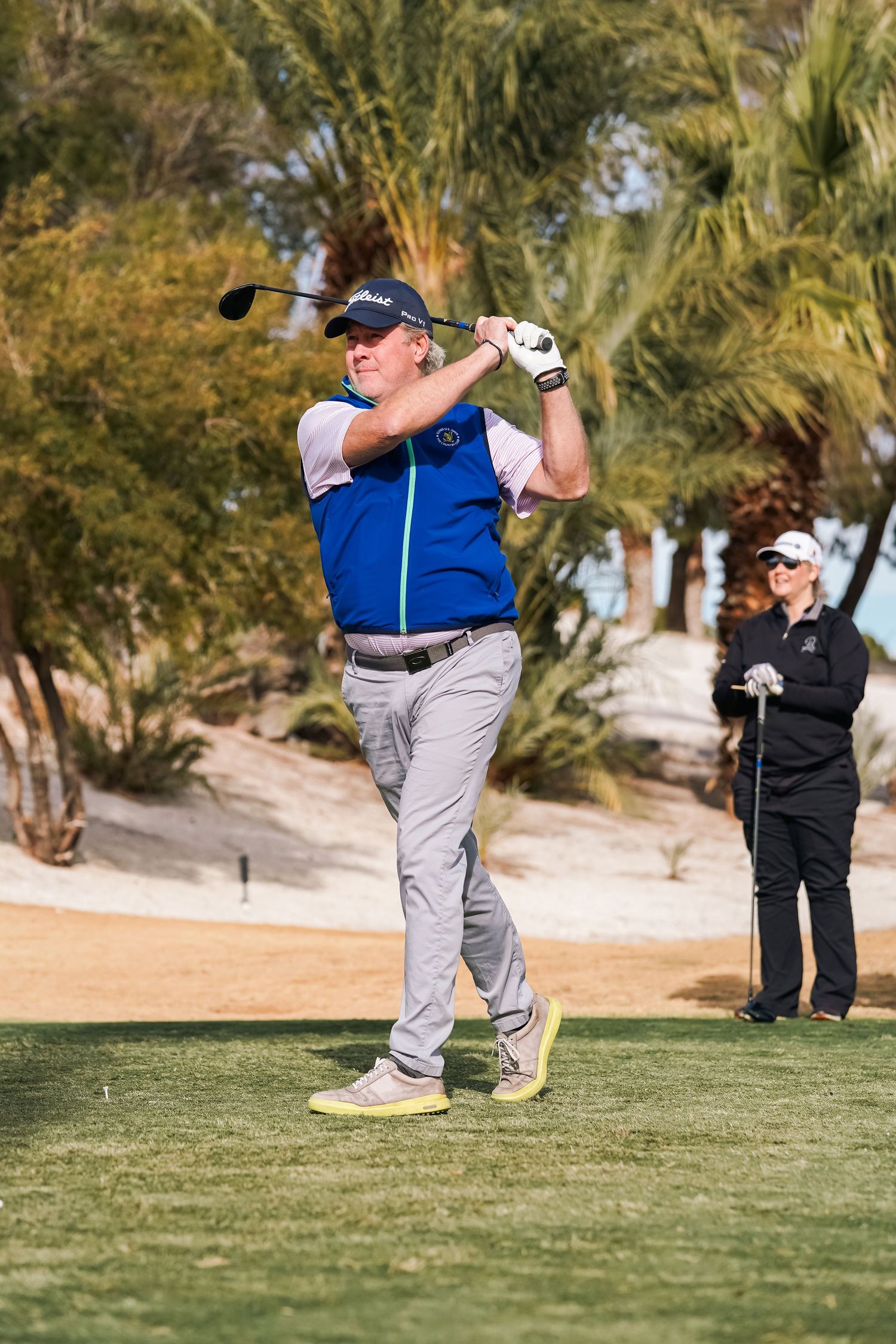 Golfer in blue vest taking a full swing while another player watches in the background.