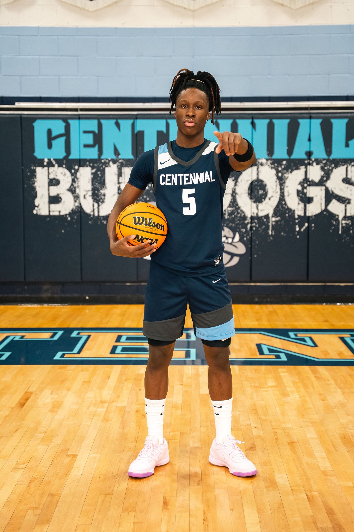 Basketball player in navy blue Centennial uniform holds orange ball on wooden court with team graphics on wall.