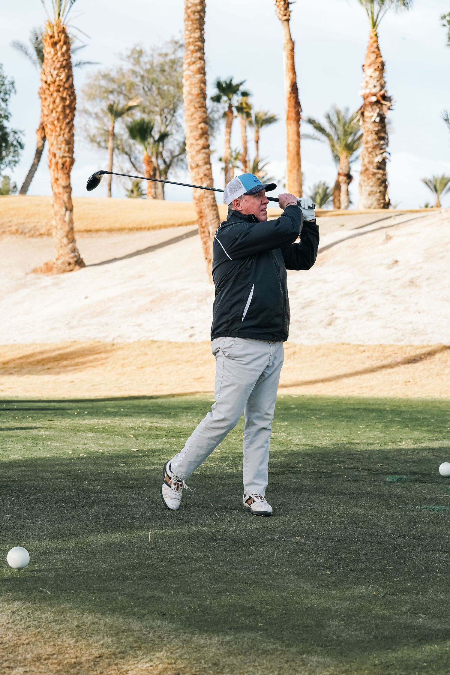 Golfer in black jacket and gray pants taking a swing at a desert golf course with palm trees in background.