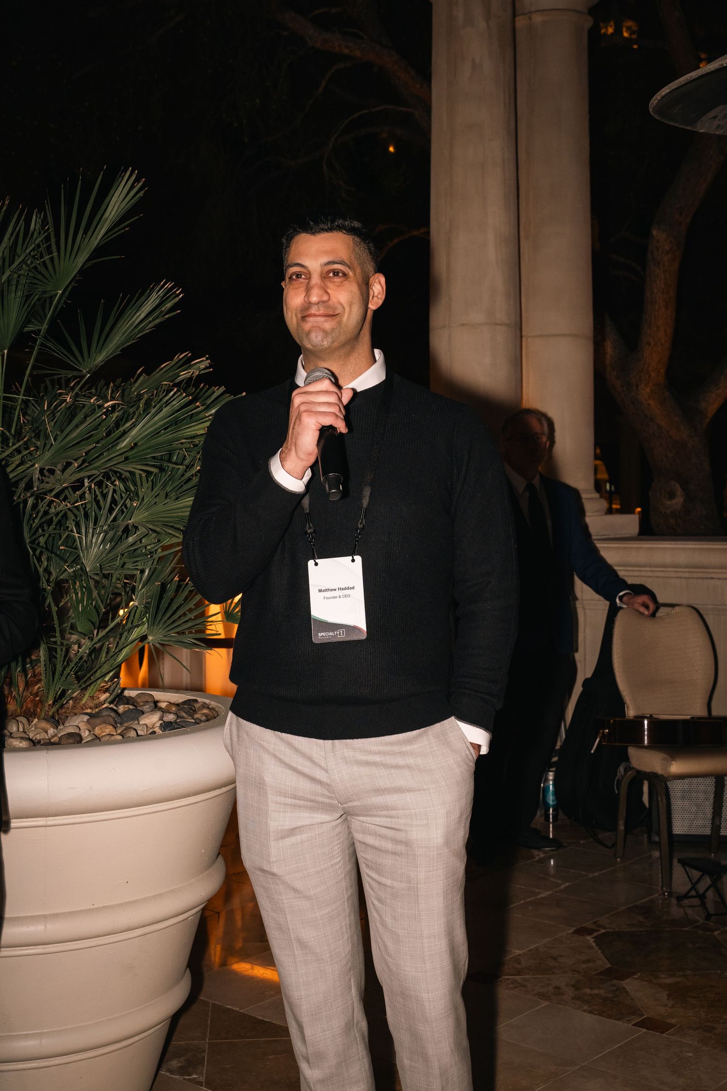 Professional attendee in business casual attire poses at an outdoor venue with palm plants.