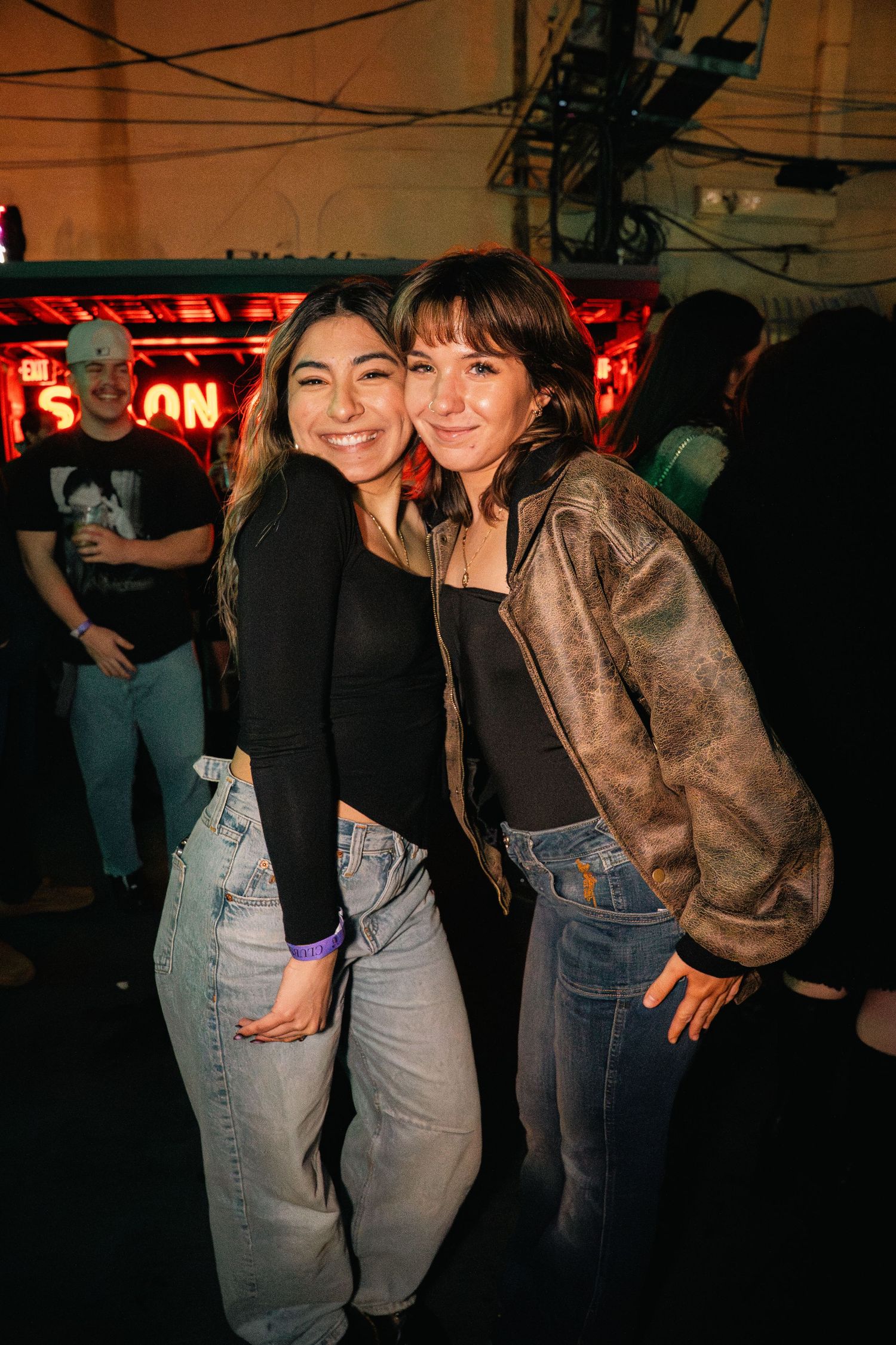 Two friends pose together at nightclub with neon signs in background.