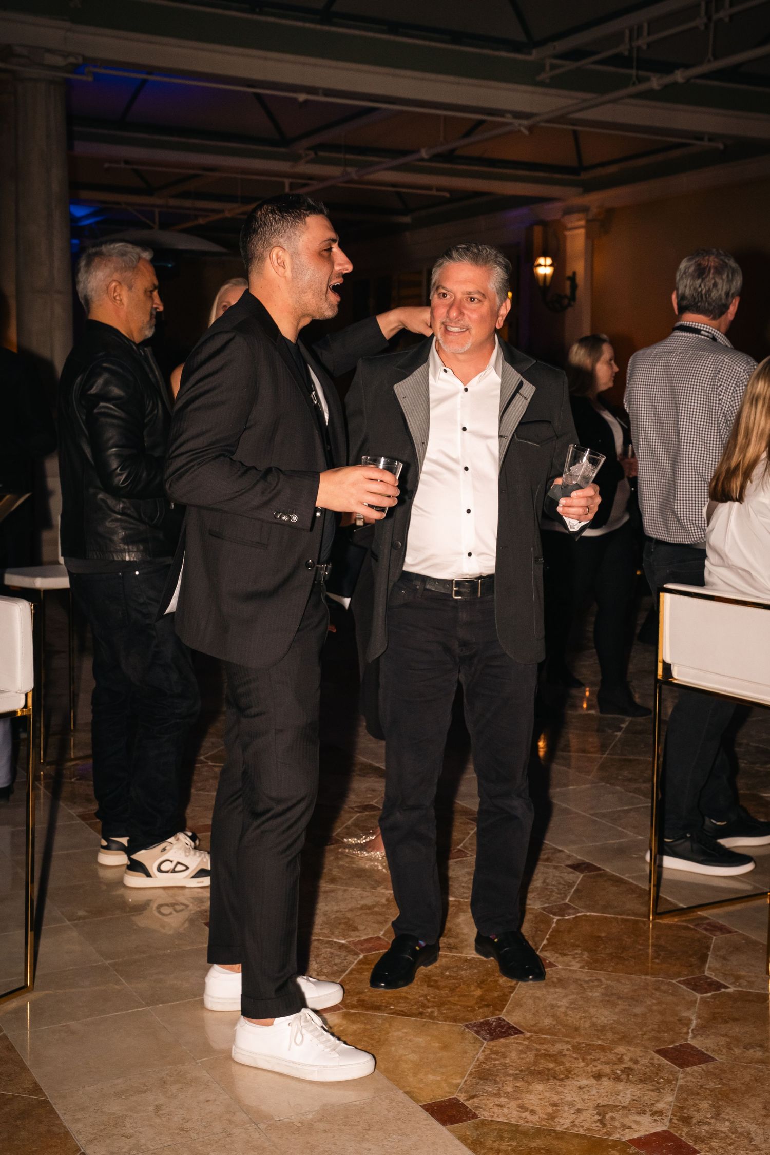 People in formal attire gather for conversation on a tiled floor at an evening reception.