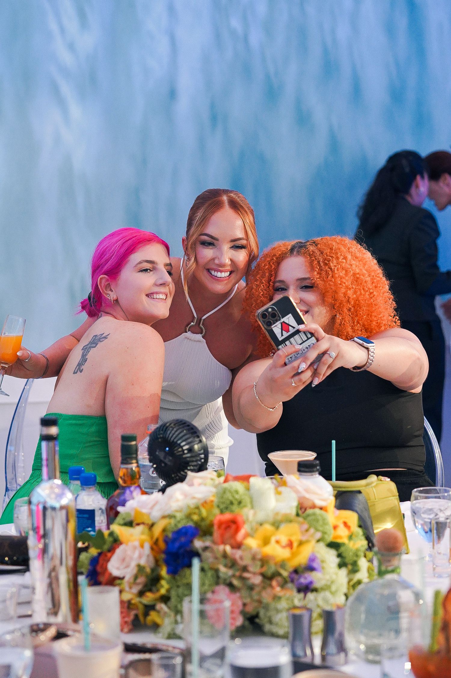 Three friends taking selfies and enjoying drinks at a celebration with colorful floral centerpieces.