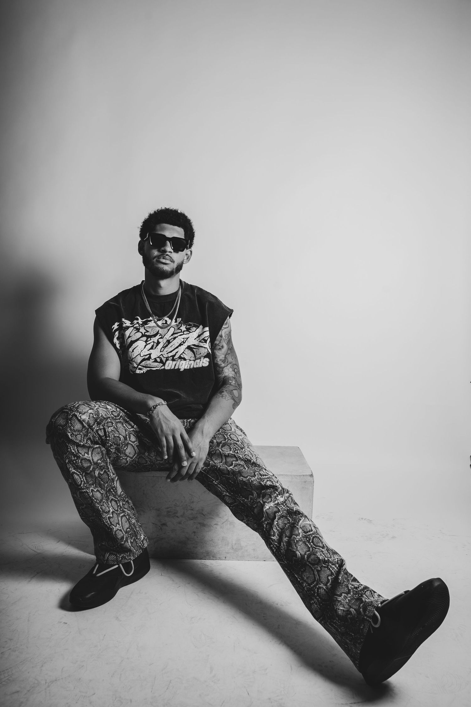 Black and white studio portrait of model in streetwear sitting casually against light background.