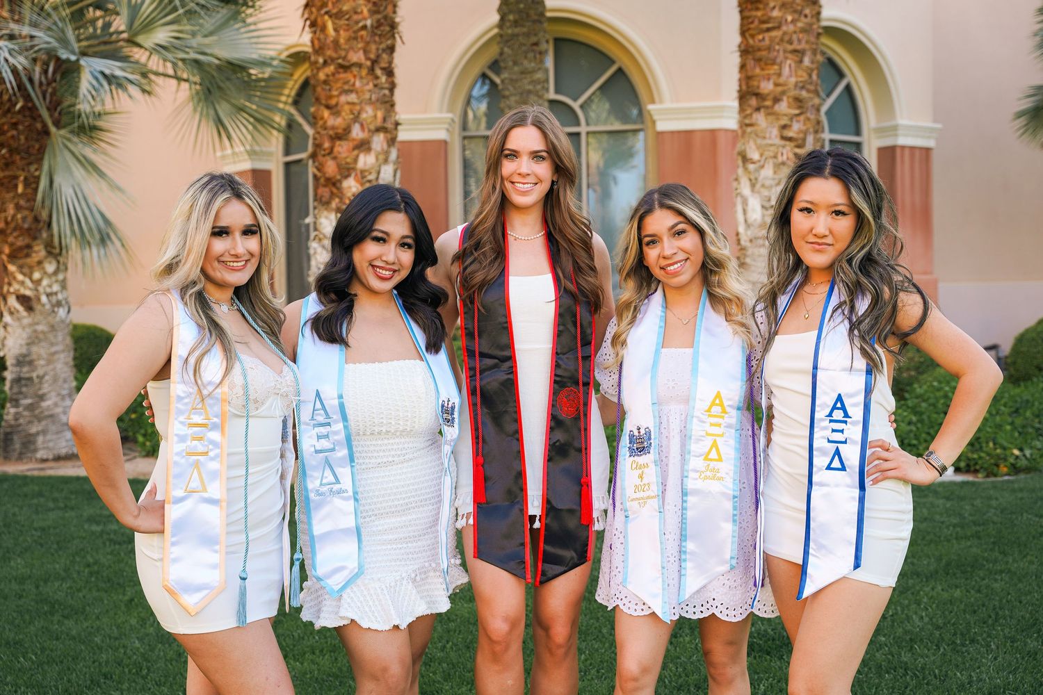 Five graduates in white outfits with graduation sashes pose together on lawn with Mediterranean building.