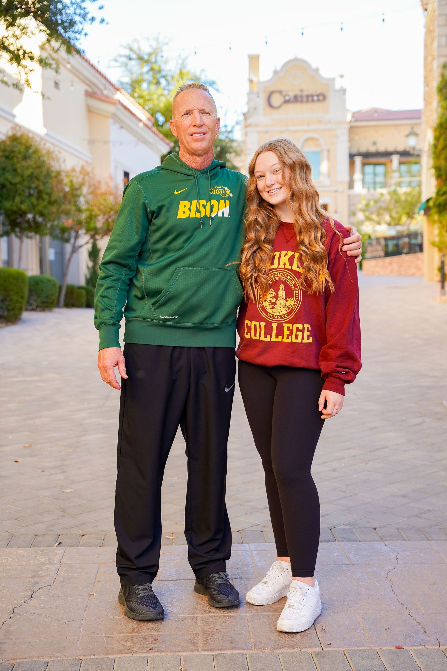 A pair in contrasting Bison green and burgundy college sweatshirts pose on a paved walkway.