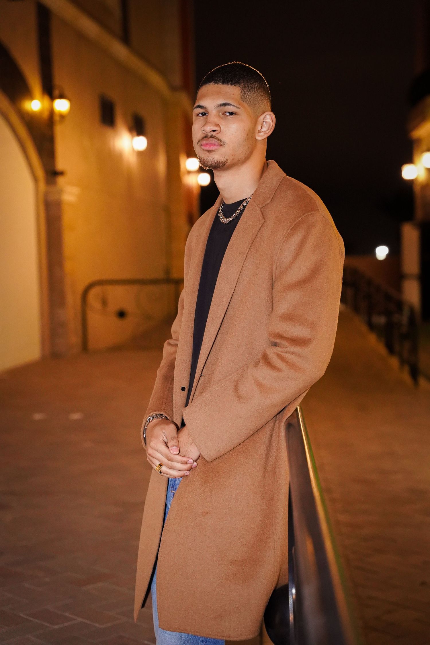 Portrait series in tan coat and black turtleneck against warmly lit architecture.