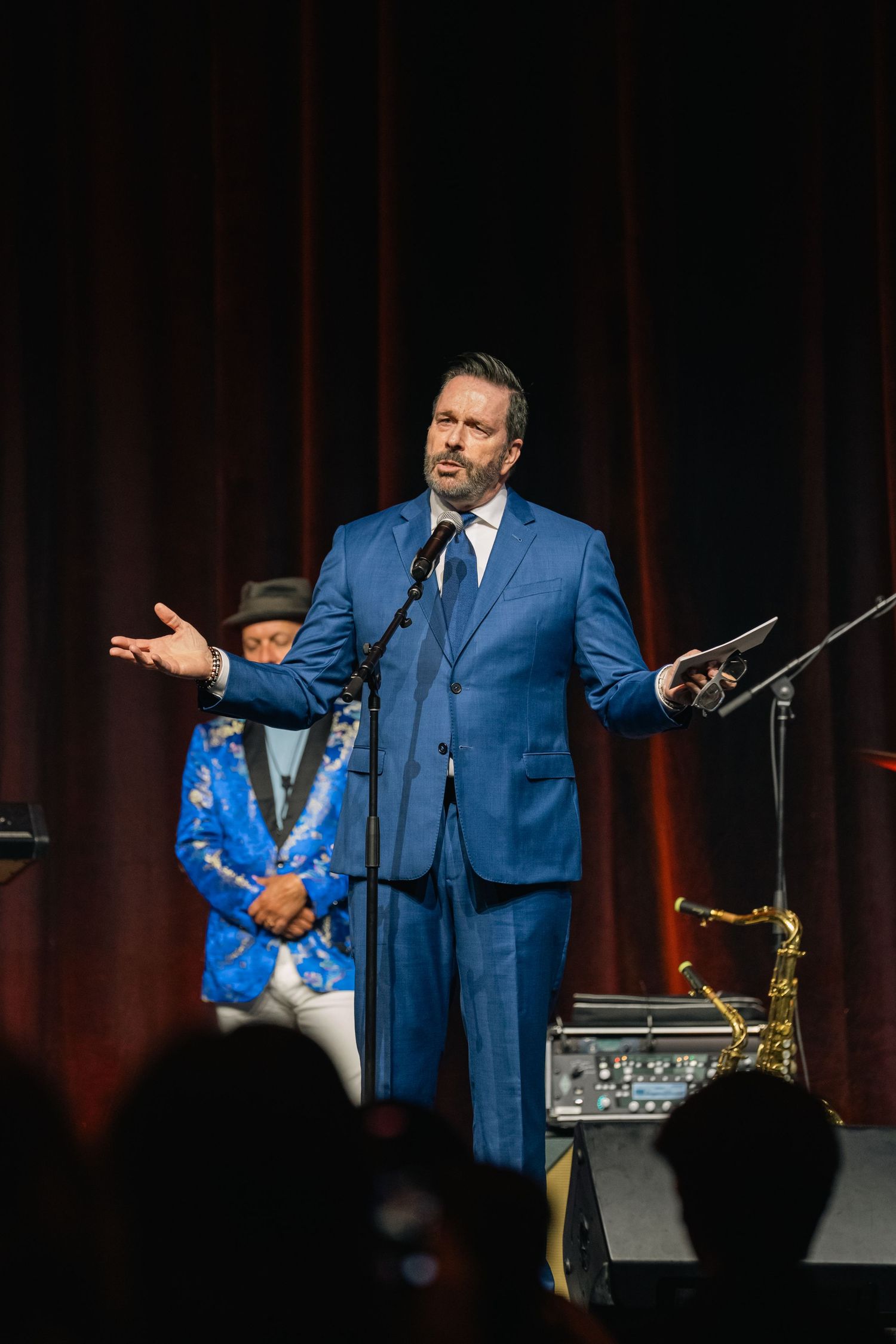 Performer in a blue suit speaking into microphone on stage with dramatic lighting.