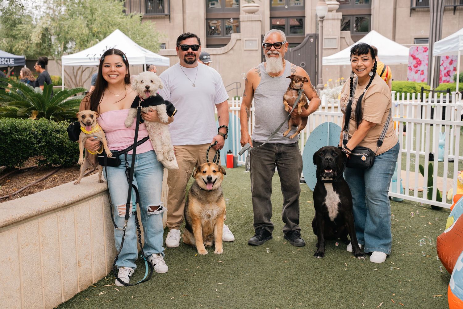 Group of dog owners and their pets posing together on artificial turf with white picket fence in background.