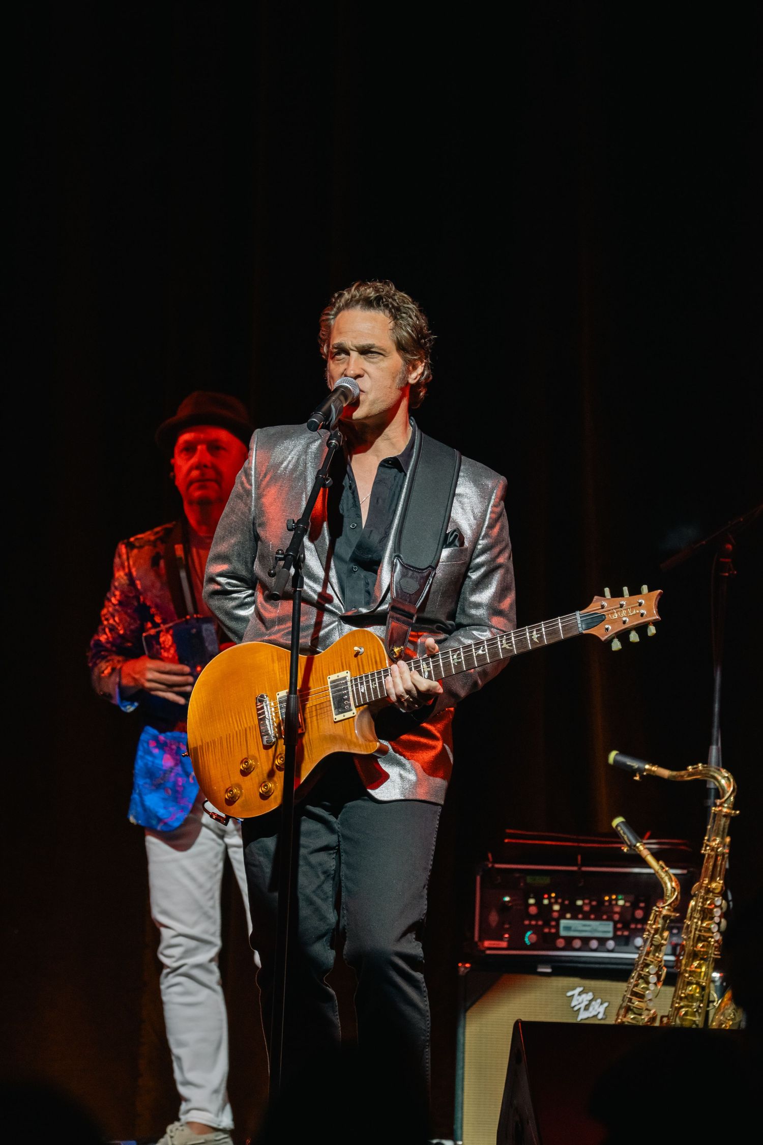 Performer singing and playing guitar on stage in silver jacket during concert performance.