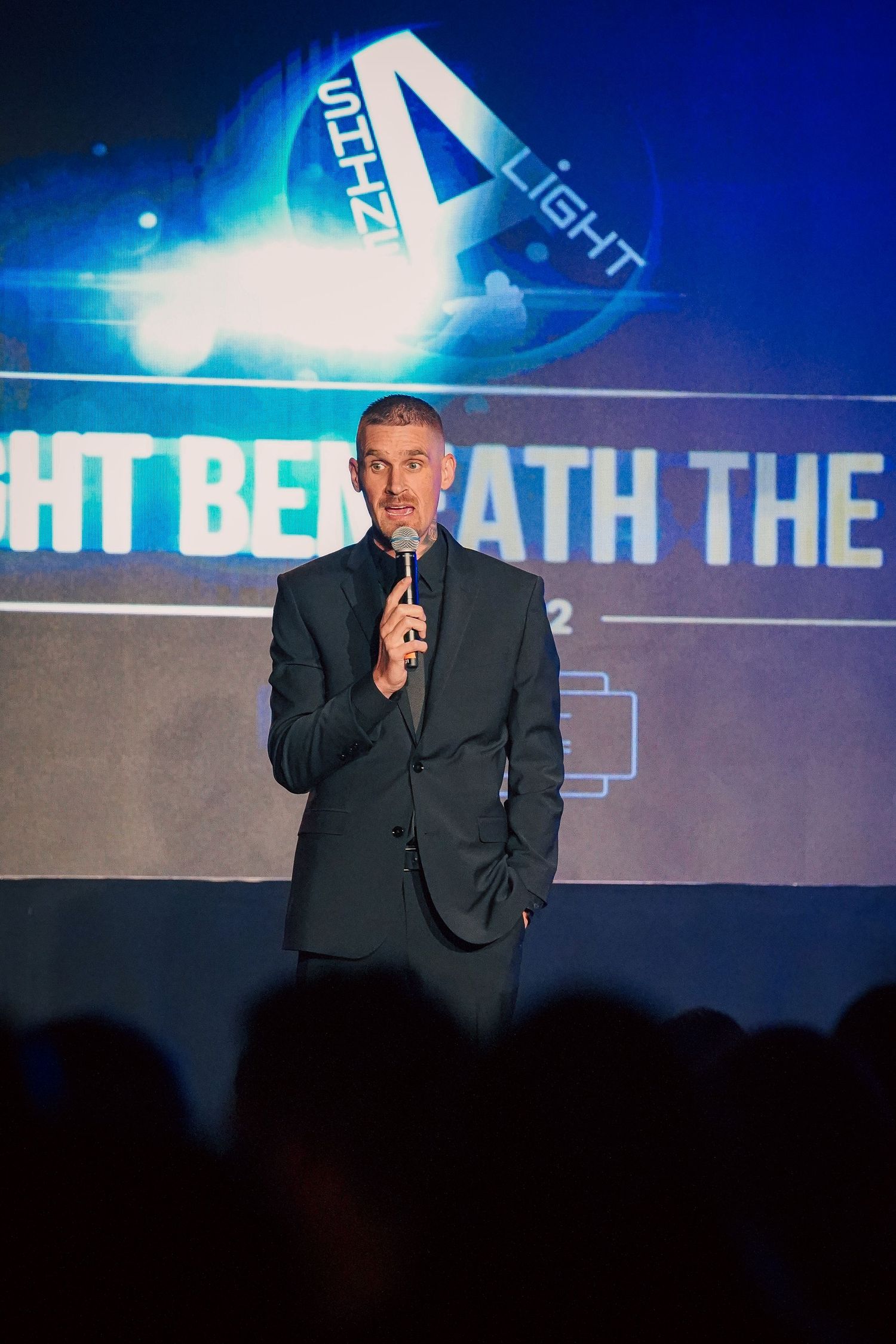 Speaker presenting on stage with blue lighting and projected text at a professional event.