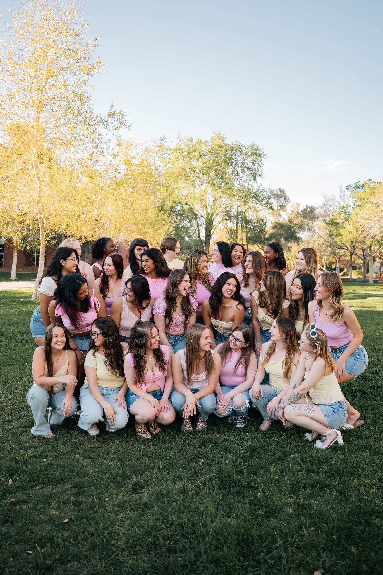 Large group of women in casual attire pose together on grass during golden hour.