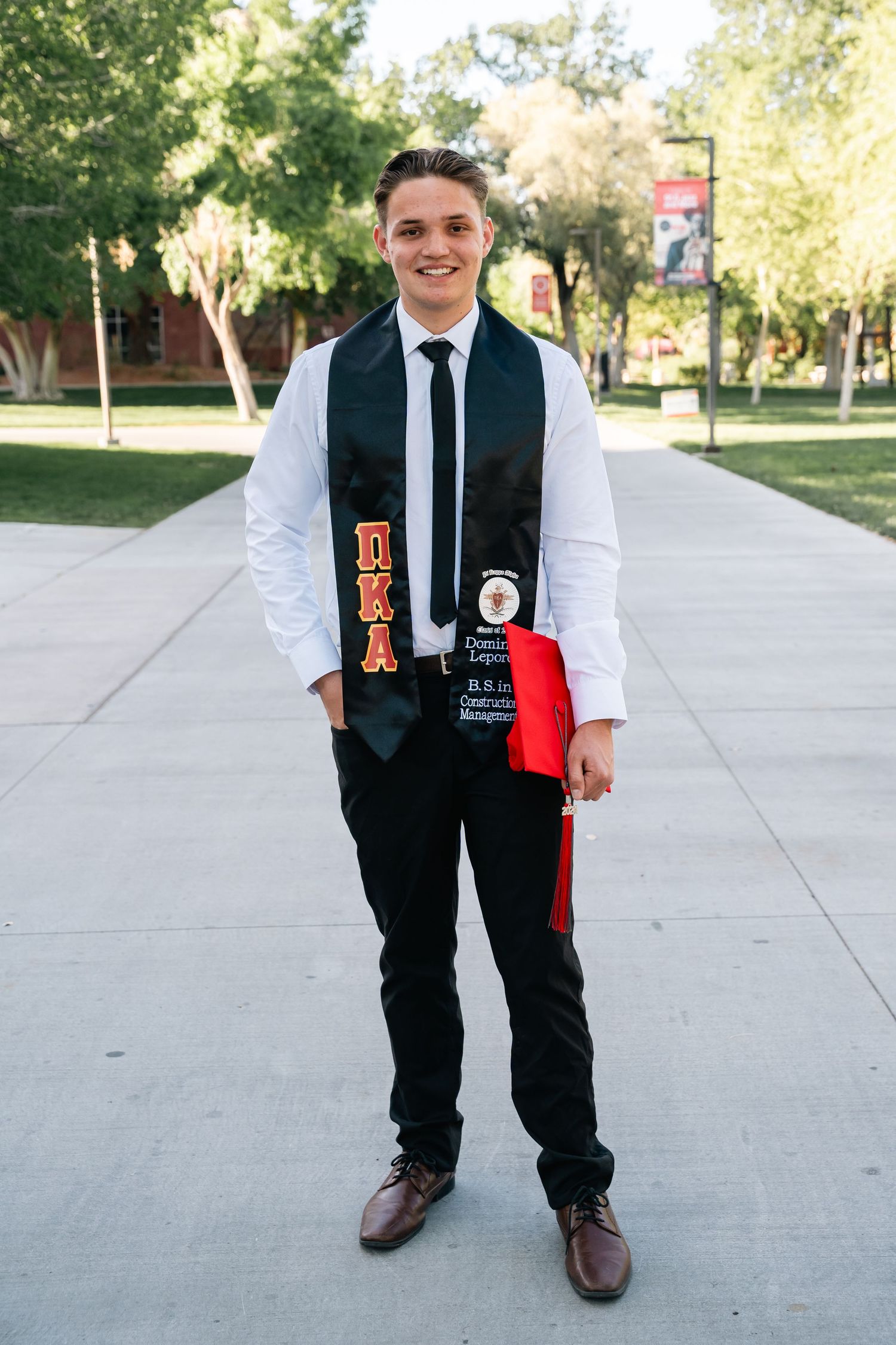 Graduate poses in formal attire with graduation stole on campus walkway.