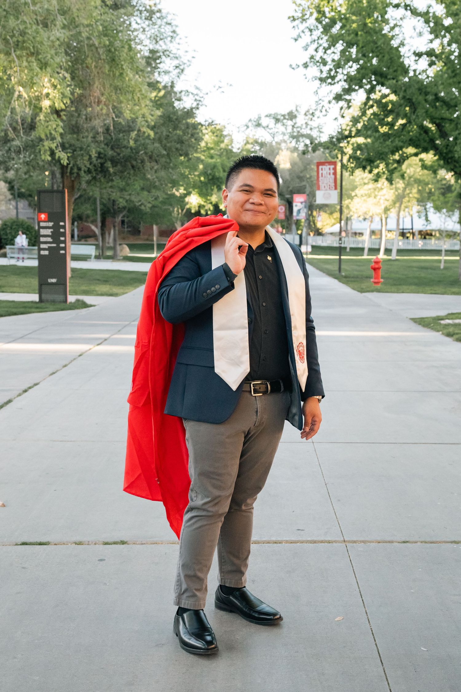 Graduate in formal attire with red graduation stole poses on campus walkway surrounded by trees.