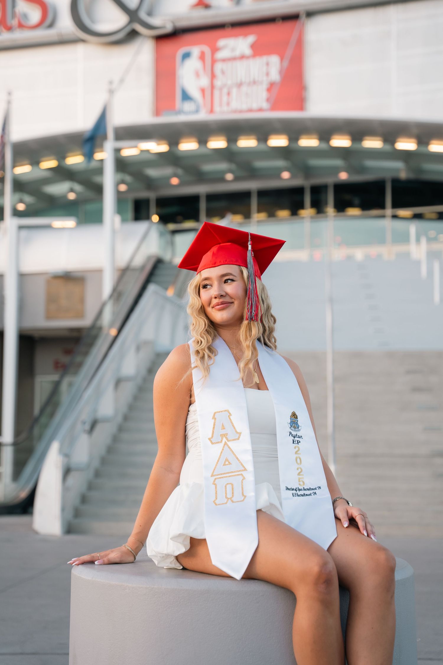 Graduate in white dress and red cap posing at UNLV campus entrance.