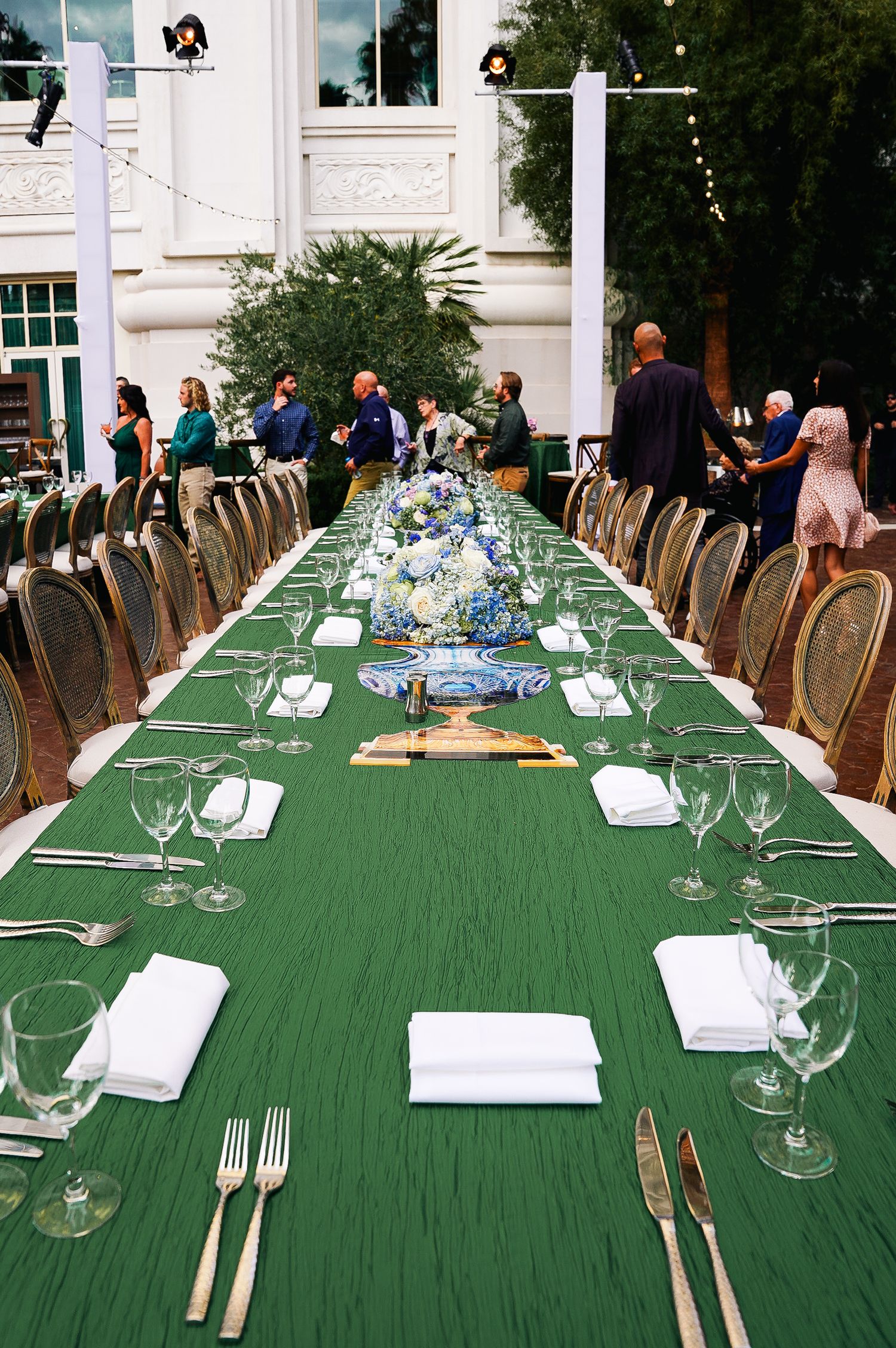 Long banquet table set with green tablecloth and formal place settings for an elegant outdoor event.
