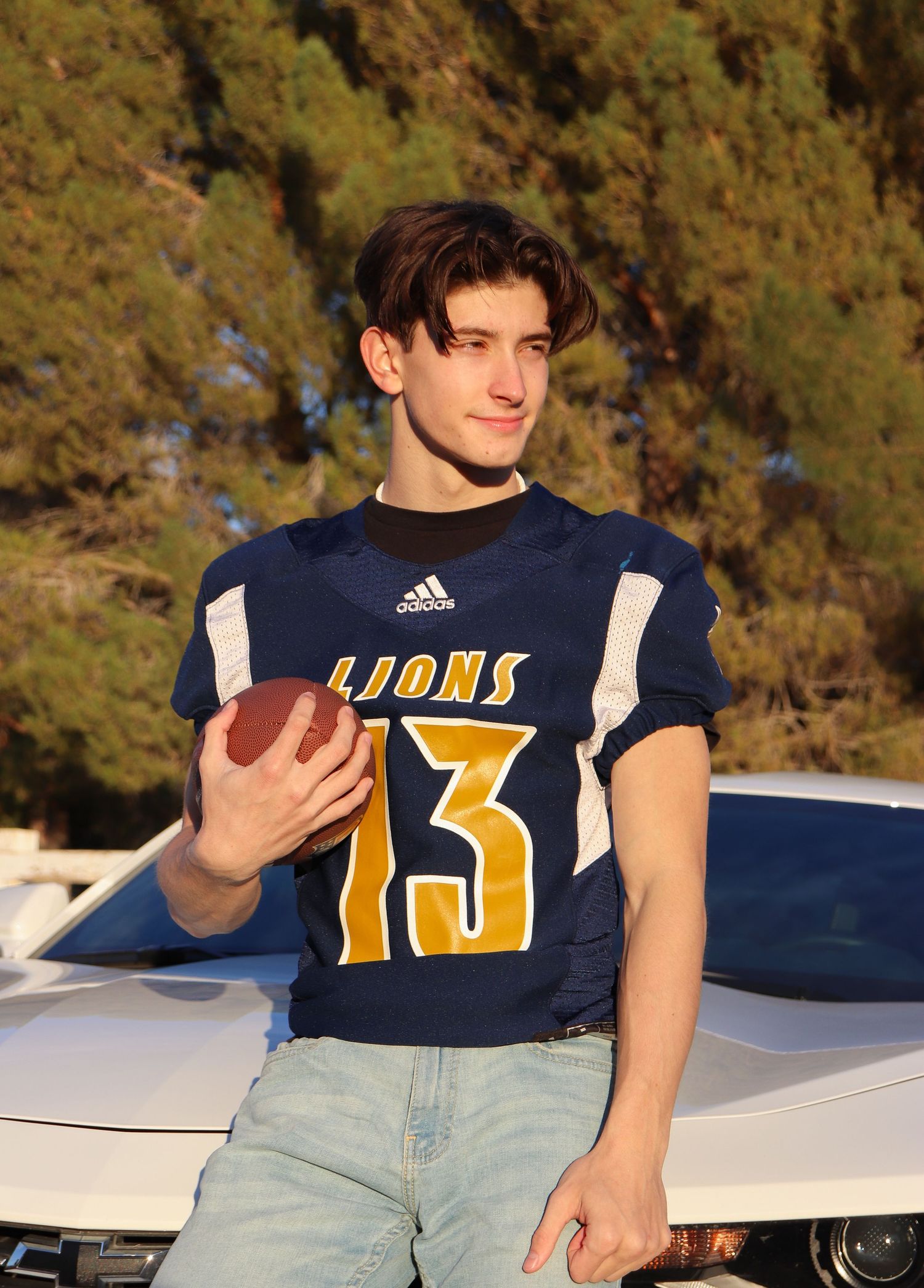 Football player in Lions #13 jersey holds ball while posing against car and trees.