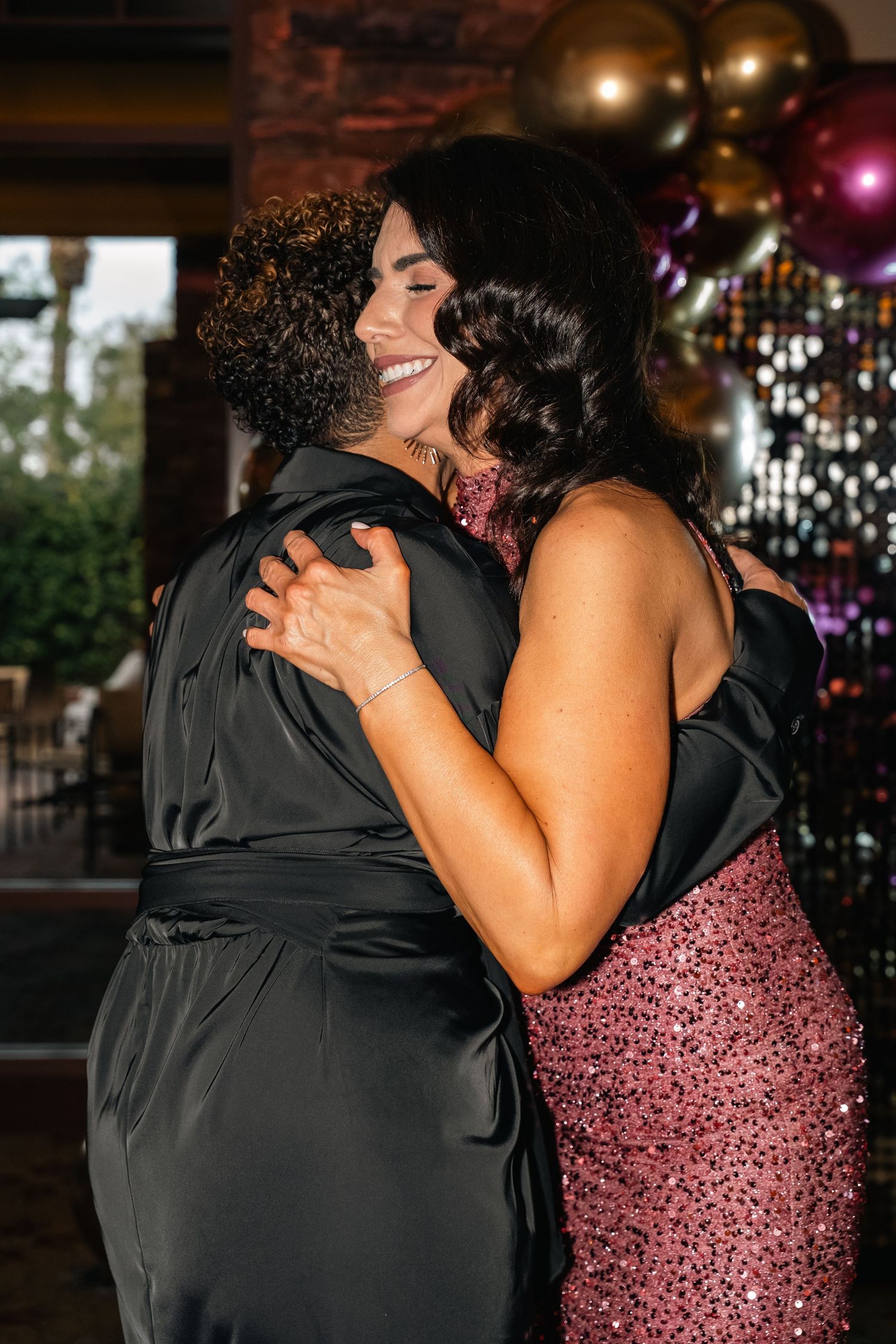 Two party guests share a warm embrace at an evening celebration with balloons in the background.
