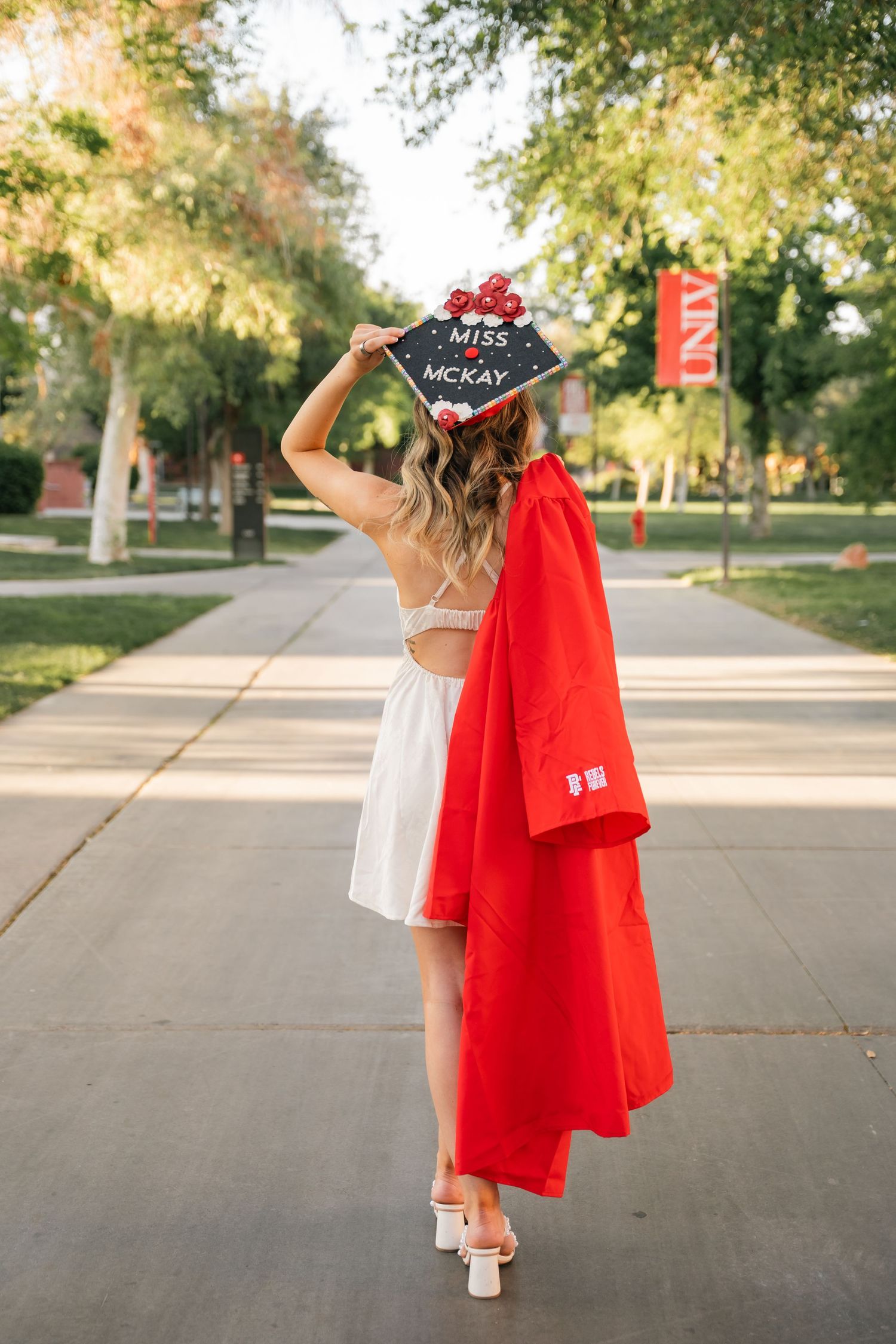 Graduate in red graduation gown shows decorated cap while walking away on tree-lined campus path.