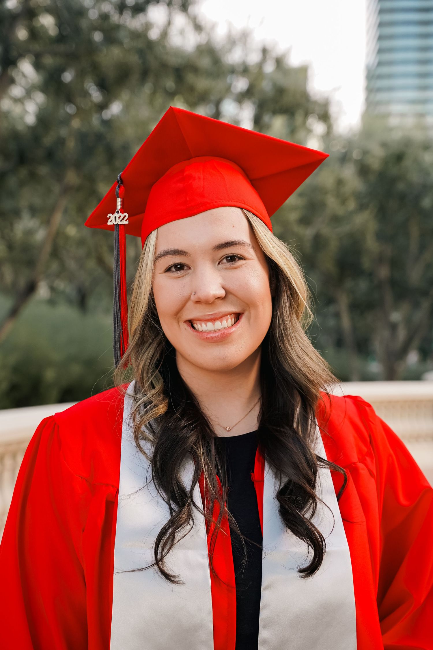 Graduate wearing red cap and gown smiles brightly against urban skyline backdrop.