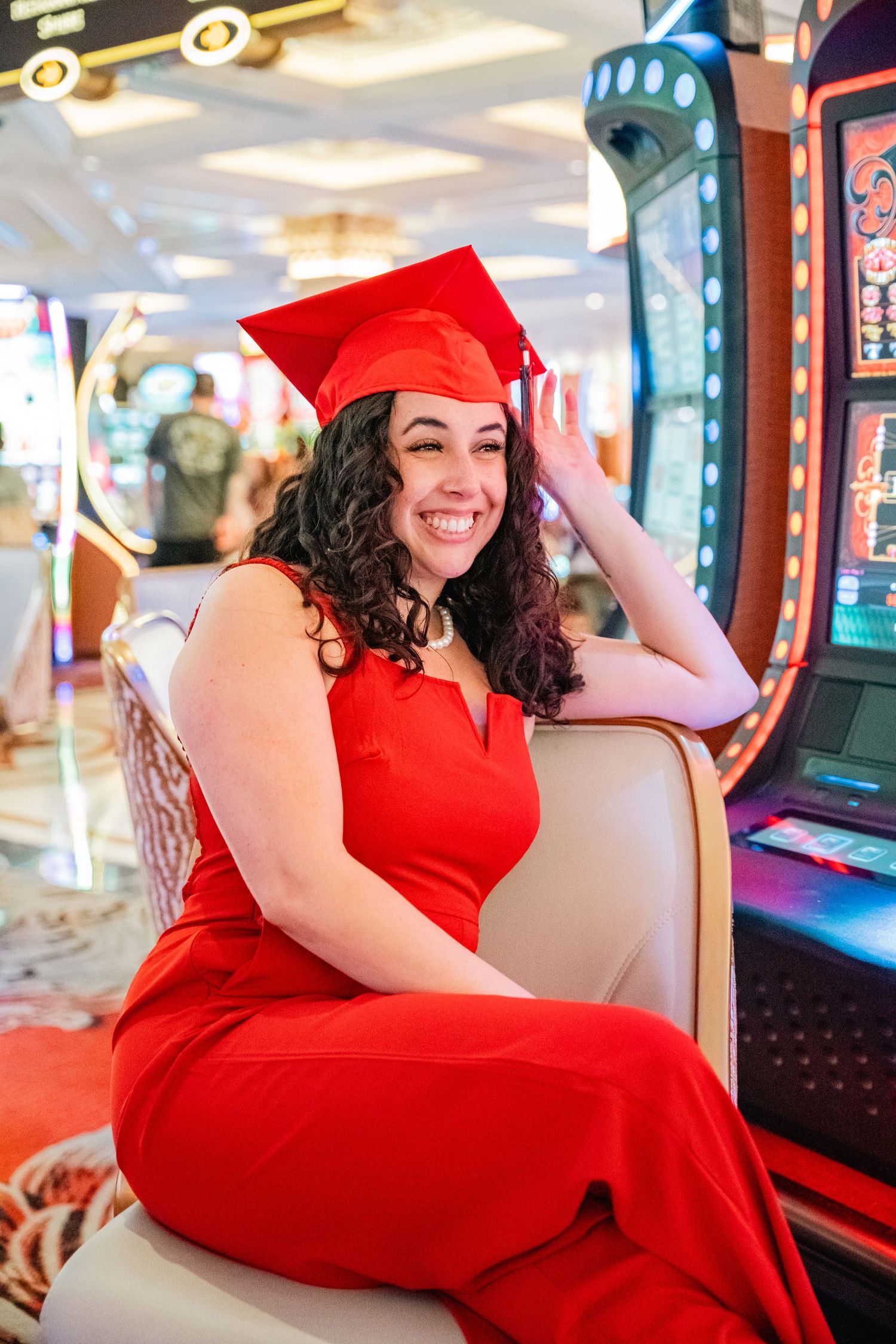 Graduate wearing red cap and gown sits by slot machines in casino setting.