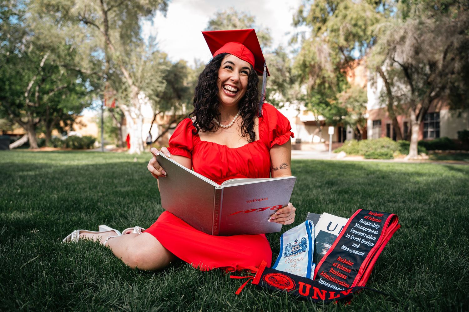 Graduate celebrating on grass wearing a red dress and cap in a sunny outdoor setting.
