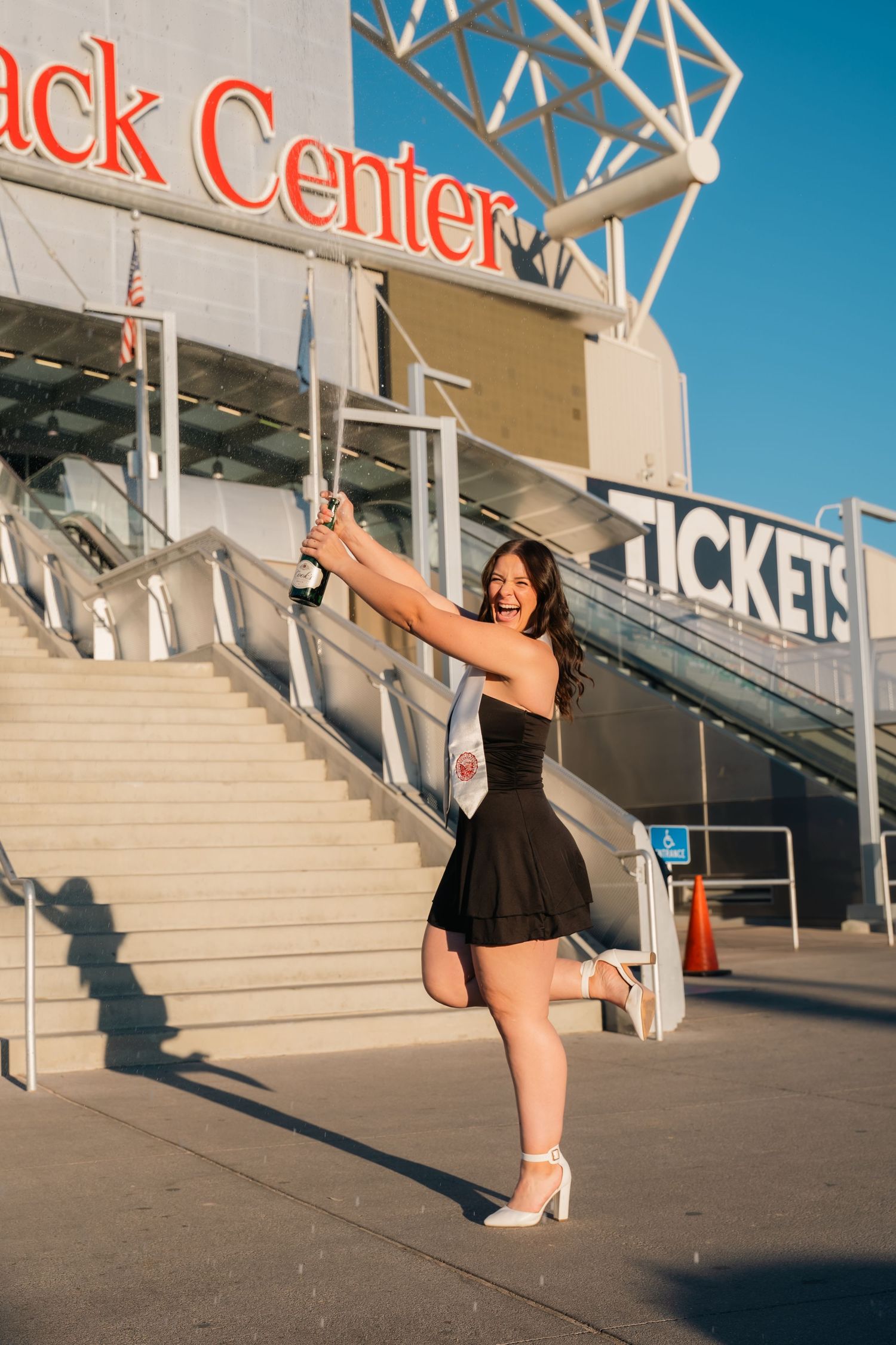 Graduate in black dress poses joyfully outside stadium ticket entrance.
