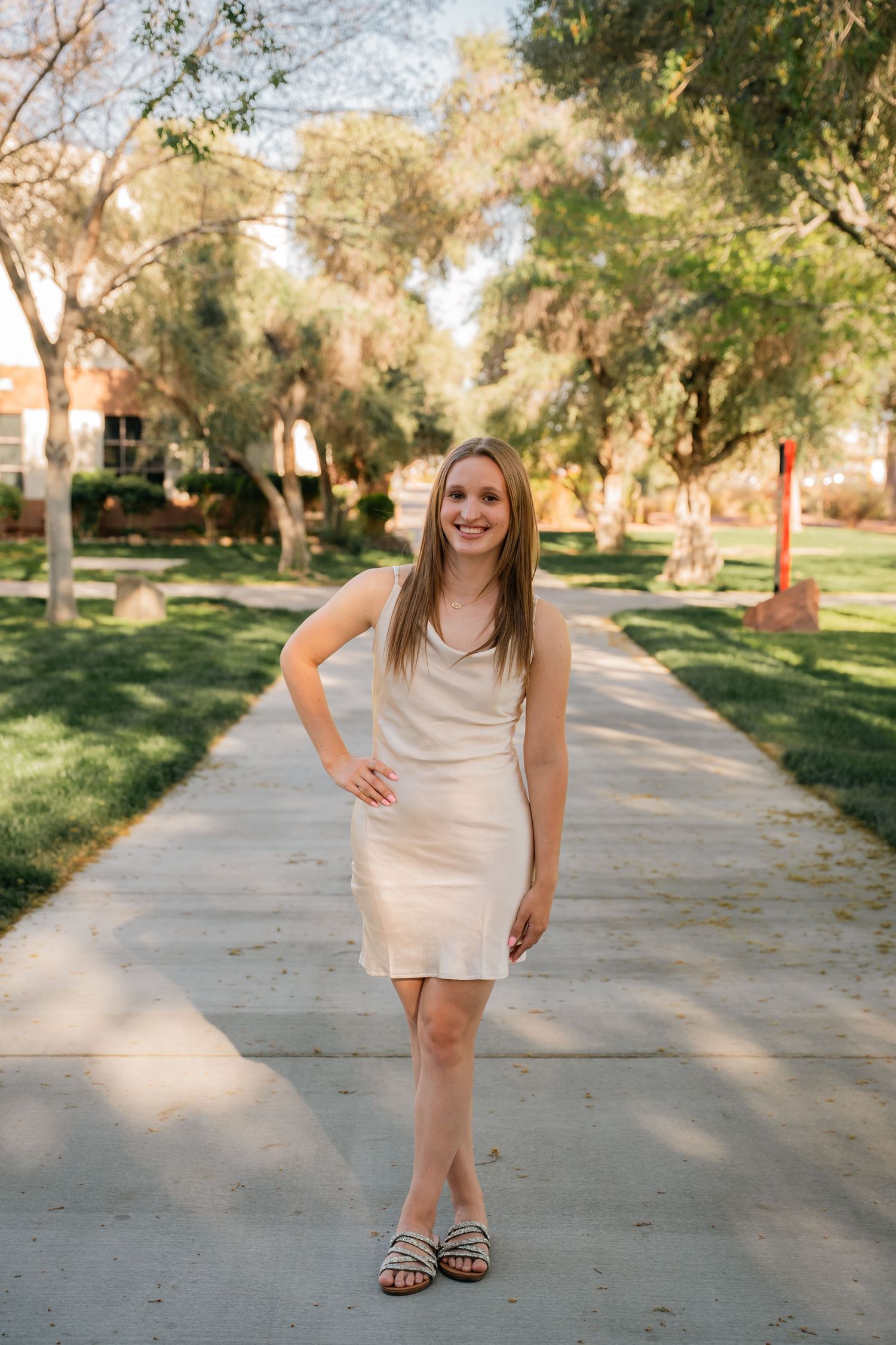 Woman in beige dress poses cheerfully on outdoor pathway with trees and grass.