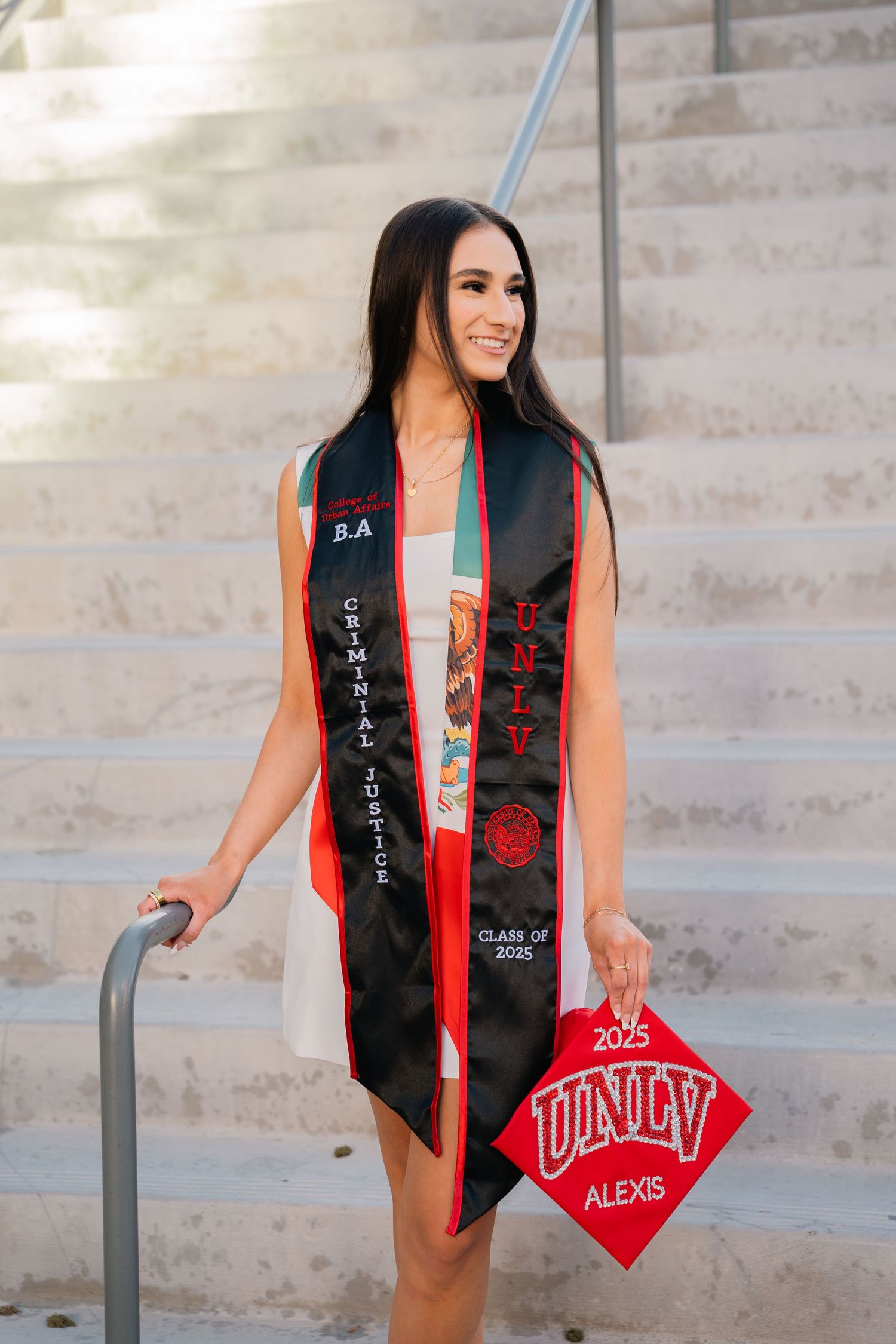Graduate with decorated cap poses on stairs with red UNLV graduation stole.