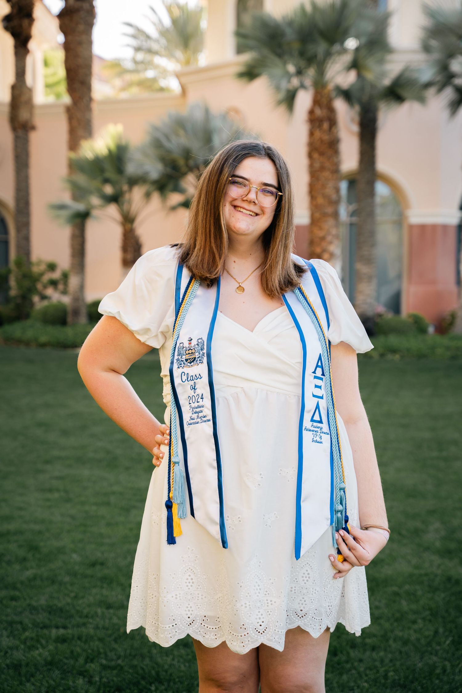 Graduate wearing white lace dress and decorated sorority stole poses by palm trees.