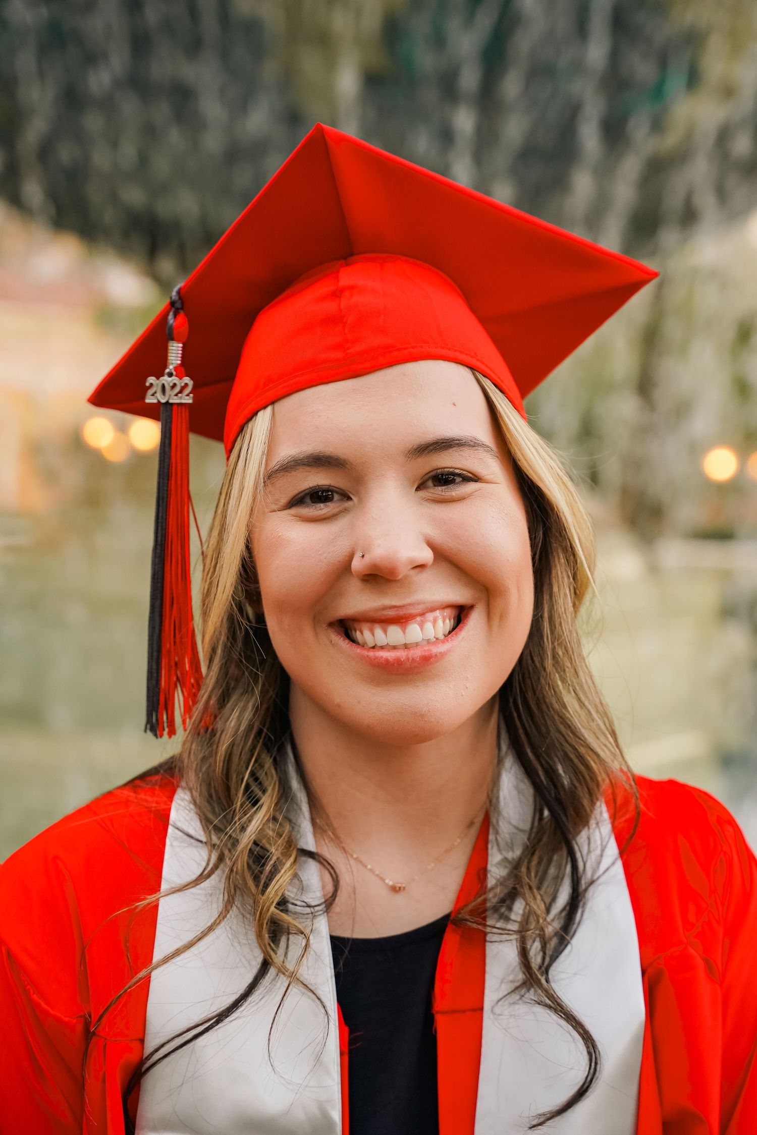 Graduate in bright red cap and gown smiles warmly against nature backdrop.
