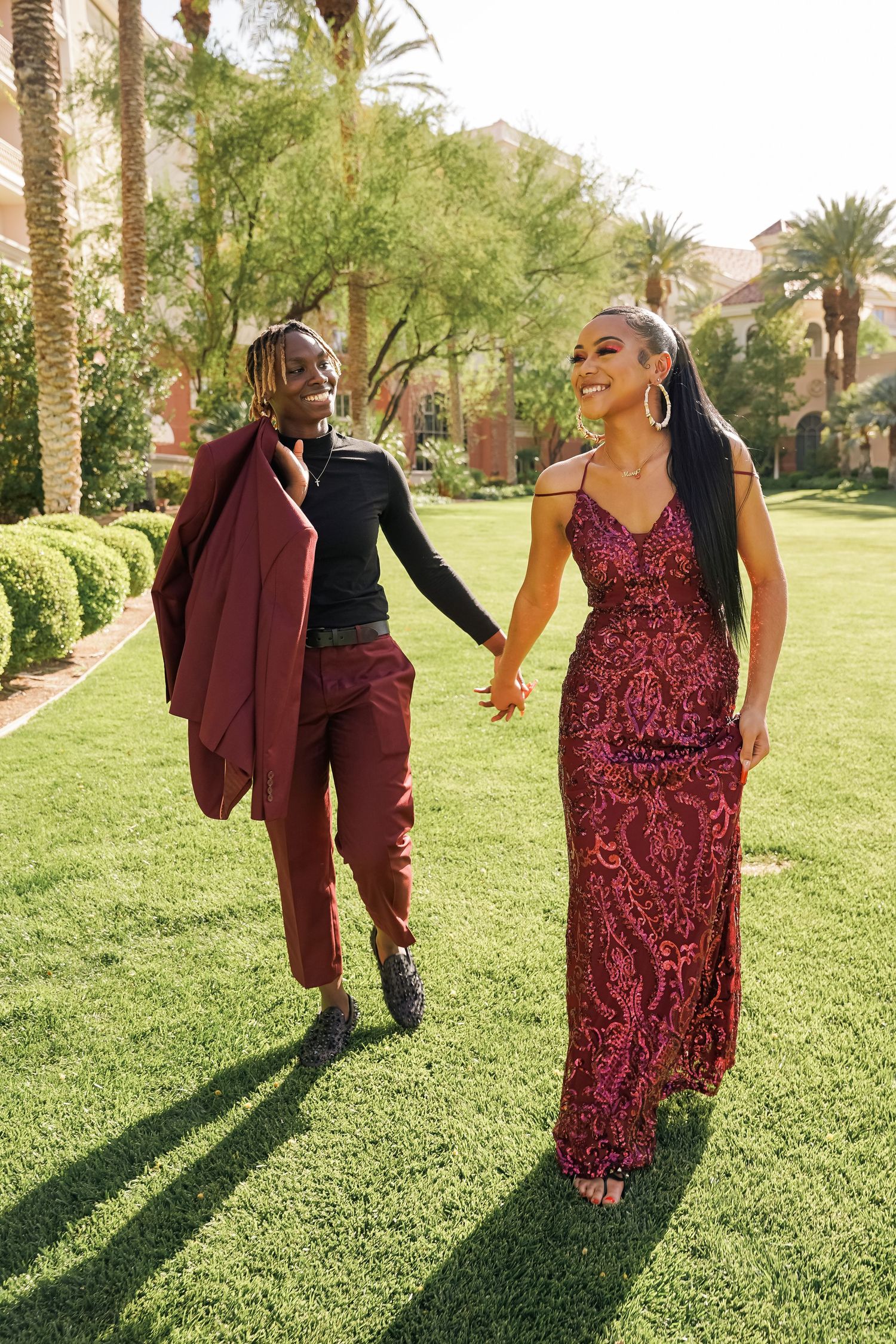 A couple in matching burgundy attire holds hands while walking through a lush garden setting.