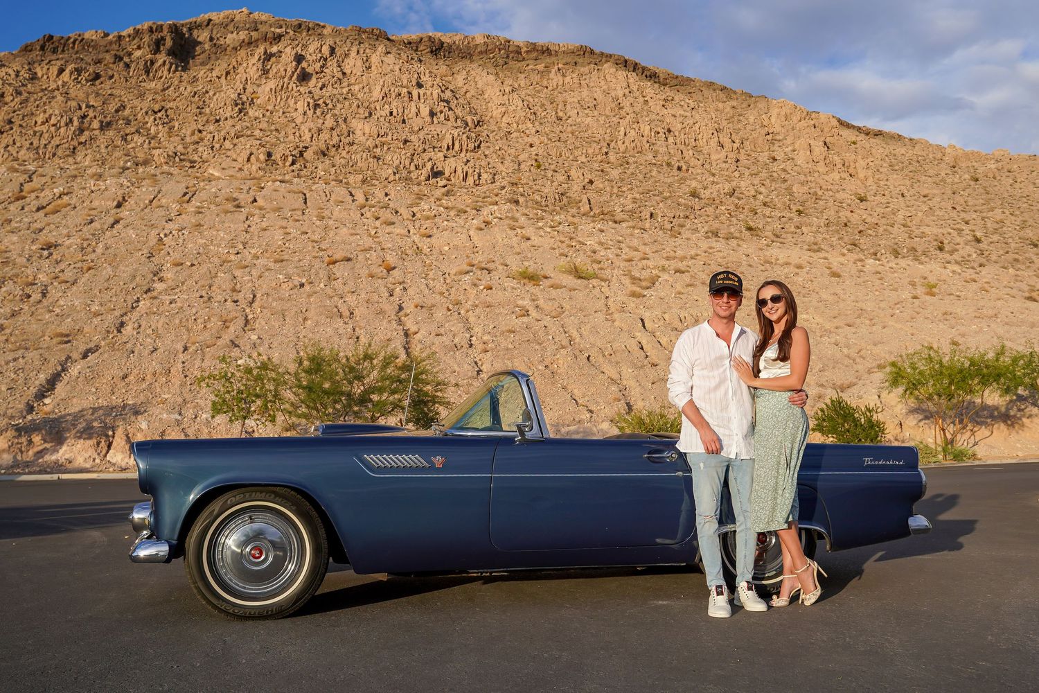 Wide panoramic shot of a couple with their vintage car against desert cliffs.