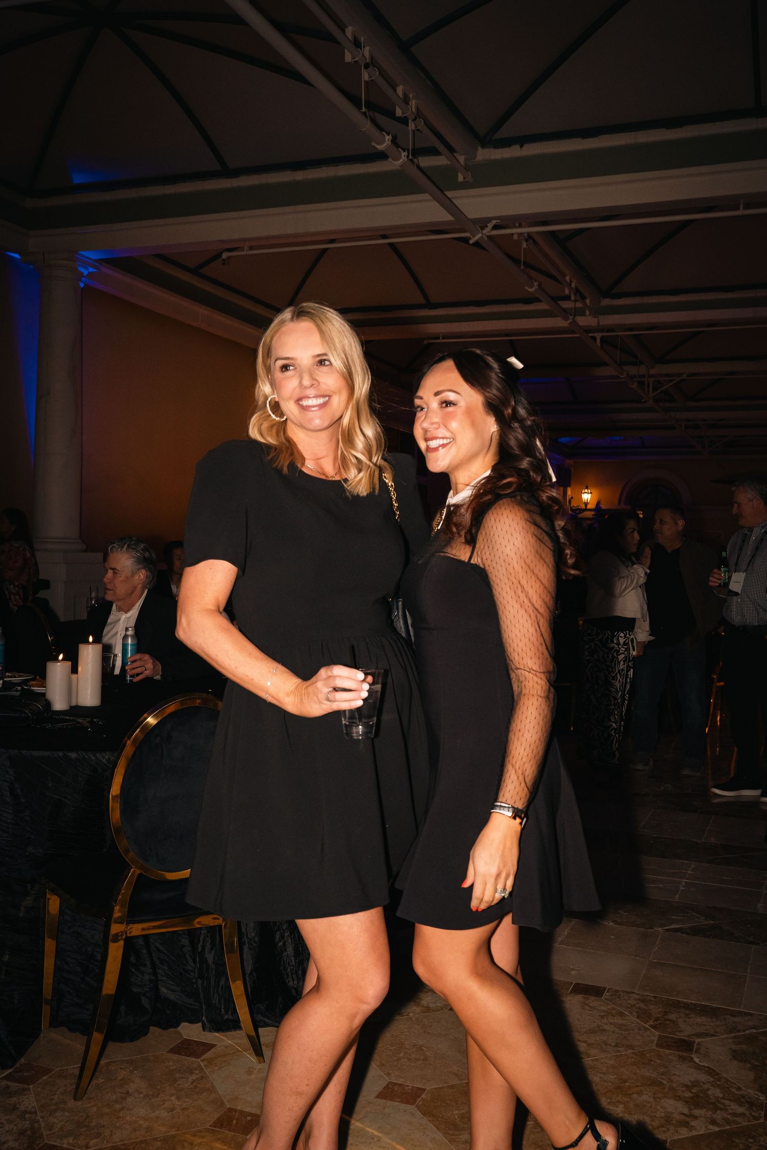 Two friends in black dresses enjoying a social gathering in elegant venue.