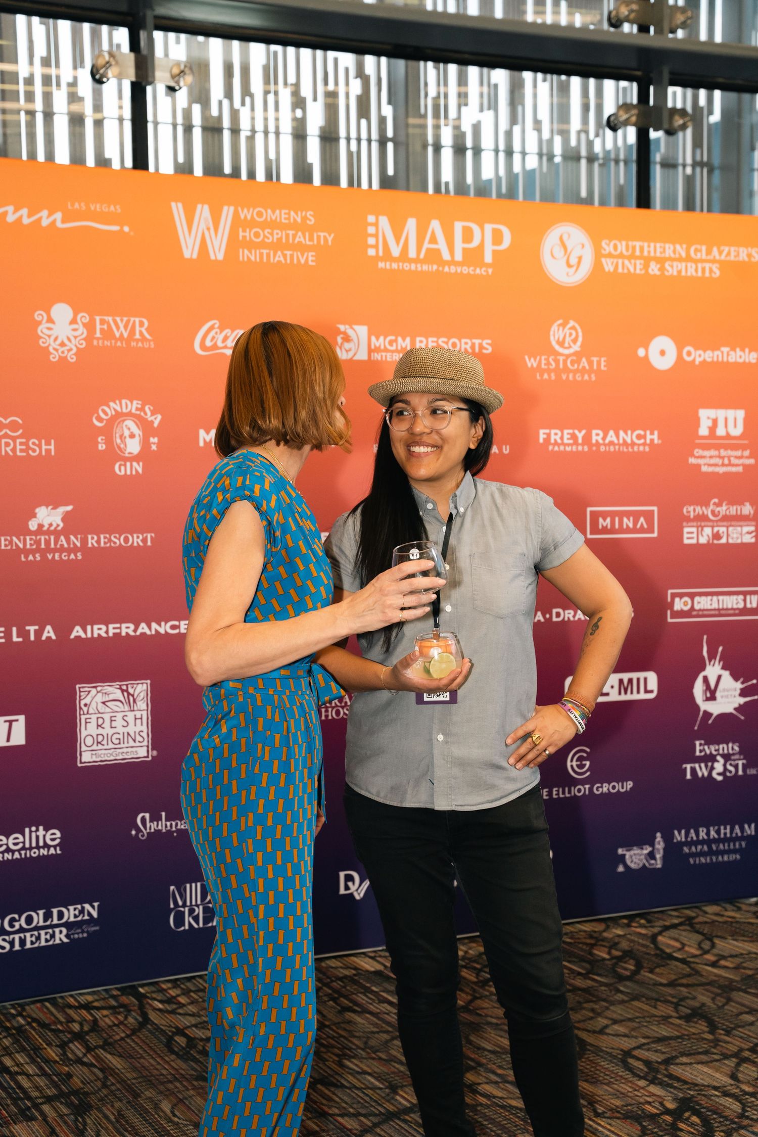 People gathering at an event with orange and purple sponsor backdrop displaying multiple company logos.