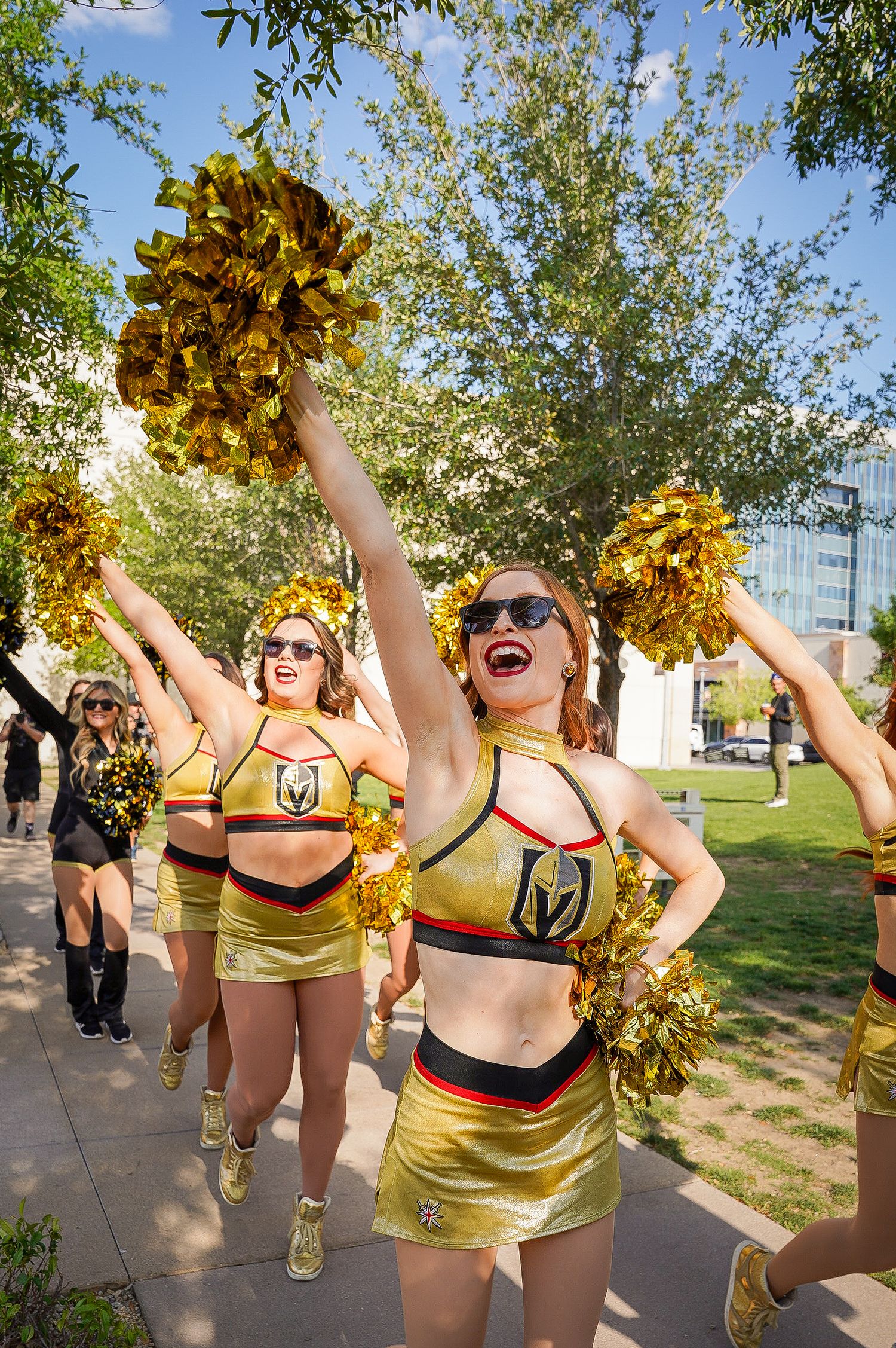 Cheerleaders in gold and black uniforms wave pom-poms enthusiastically at an outdoor event.