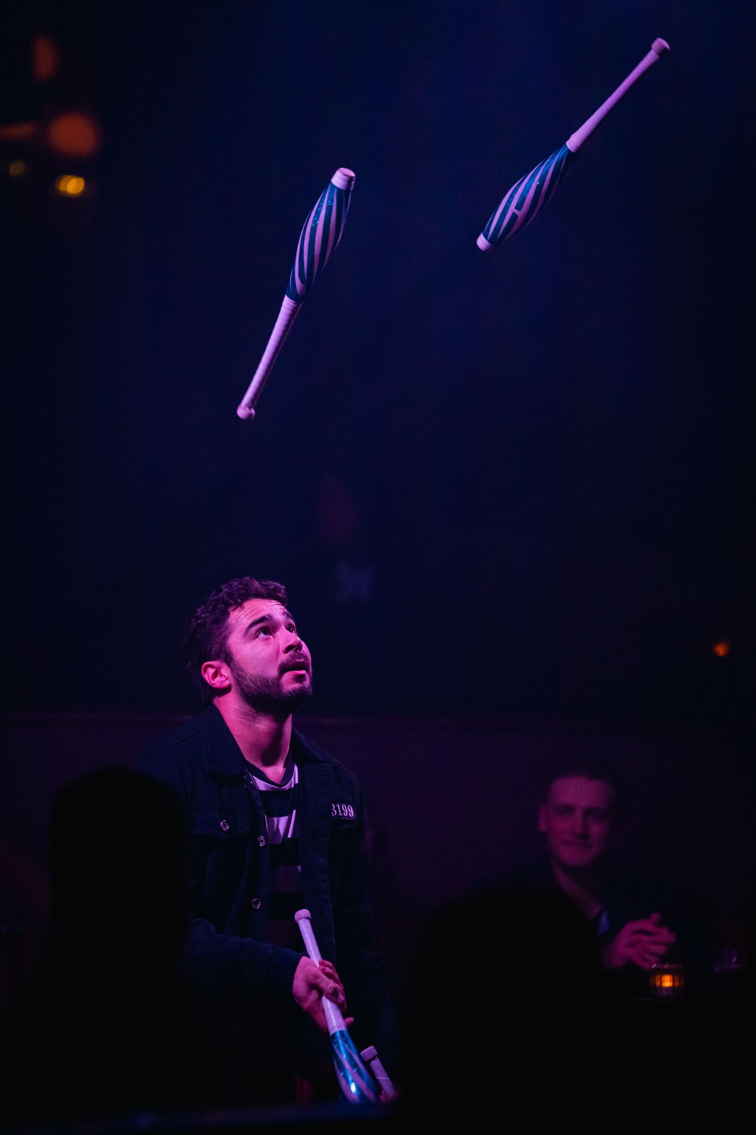 Performer juggling clubs under purple stage lighting during show.