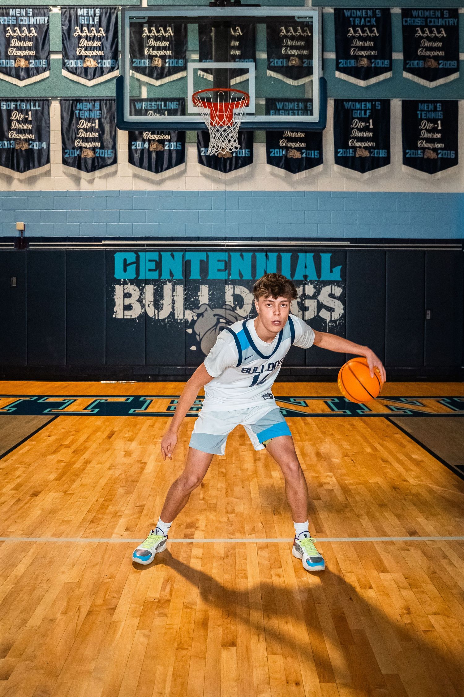 Basketball player in white Centennial Bulldogs uniform dribbles on wooden court with championship banners displayed.