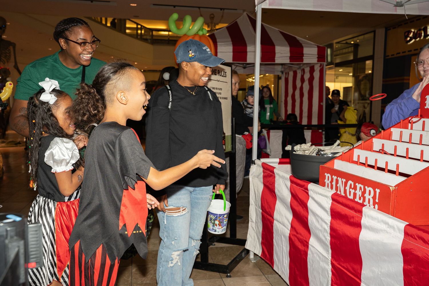 Costumed guests visit a red and white striped carnival booth at an indoor Halloween event.