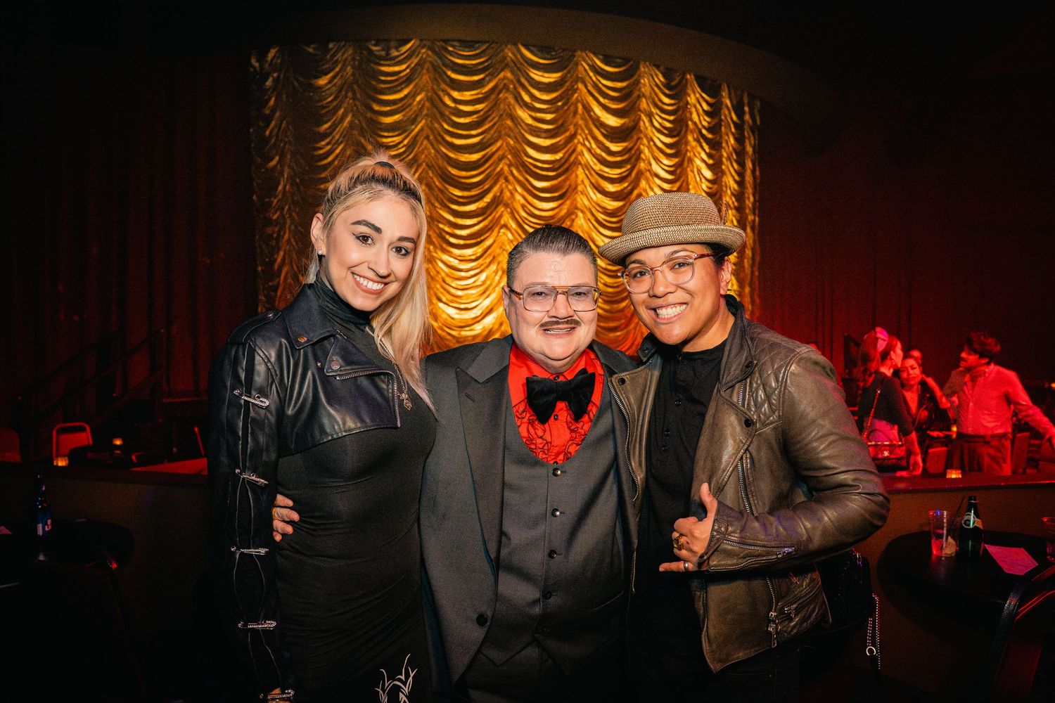 Three people pose together in front of a golden draped curtain at an entertainment venue.