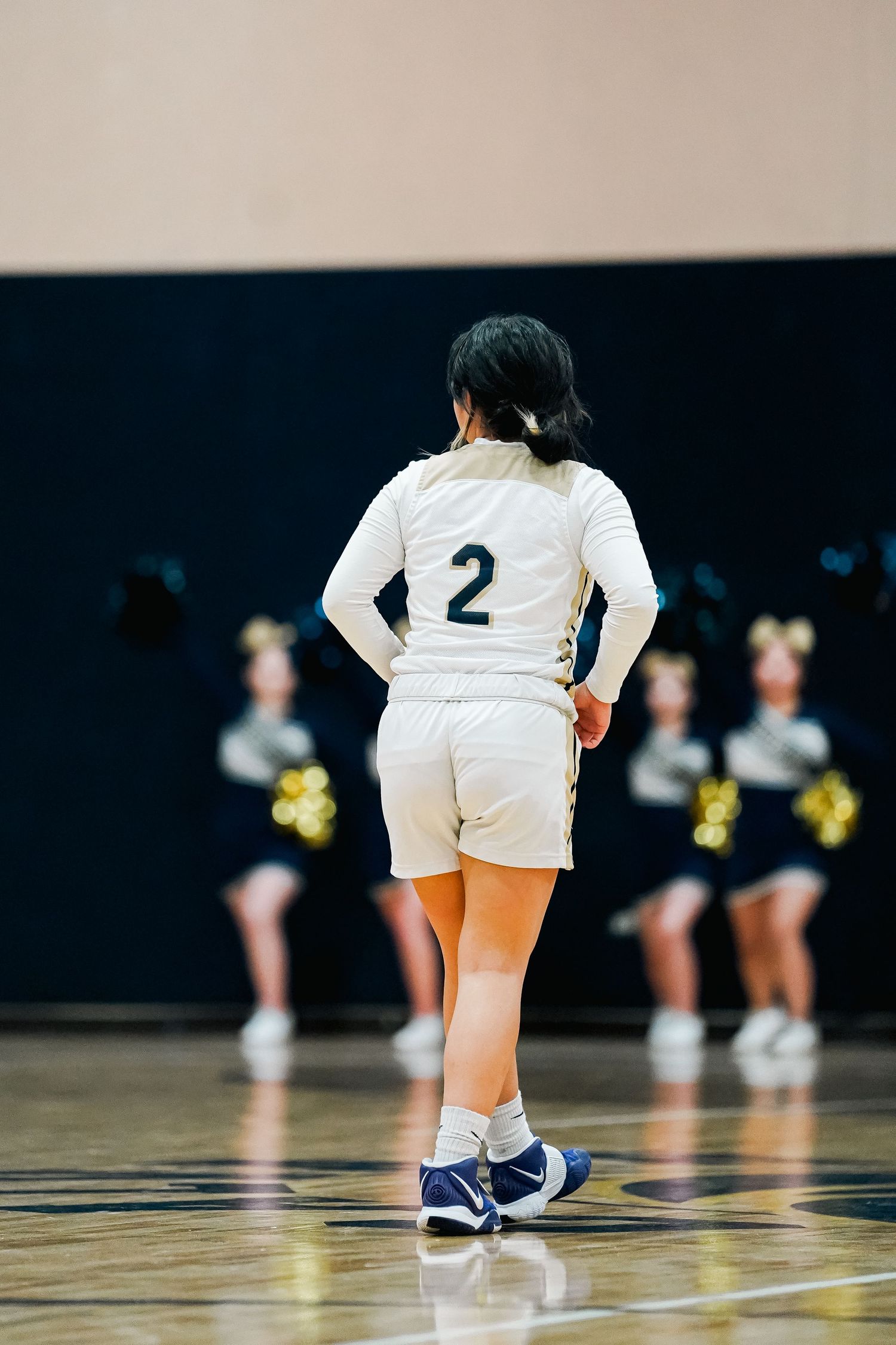 Basketball player in white uniform with number 2 standing on court with cheerleaders in background.