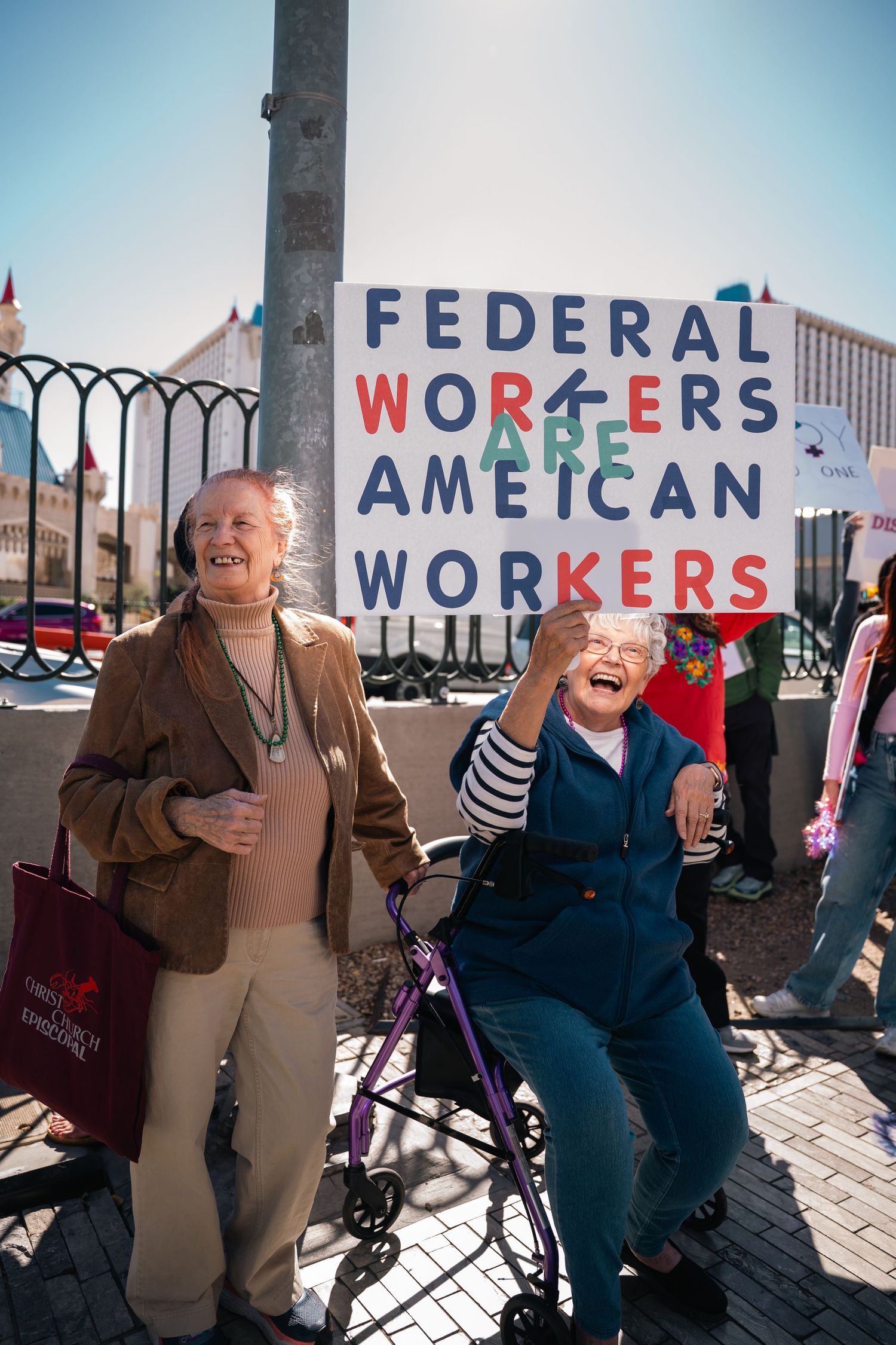 Two people laughing next to protest sign about federal workers on sunny day.