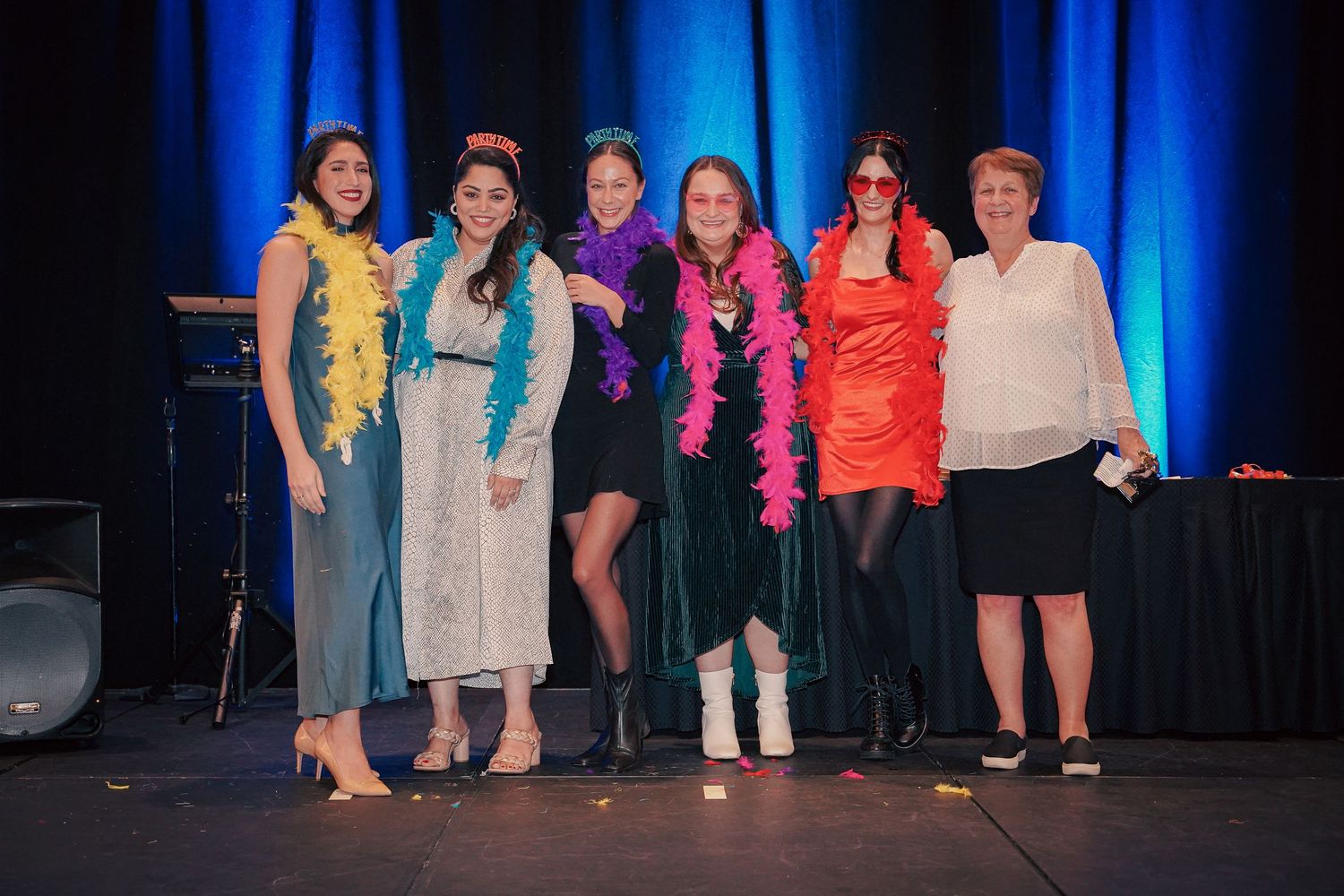 Group of performers pose together on stage wearing colorful outfits with blue backdrop.