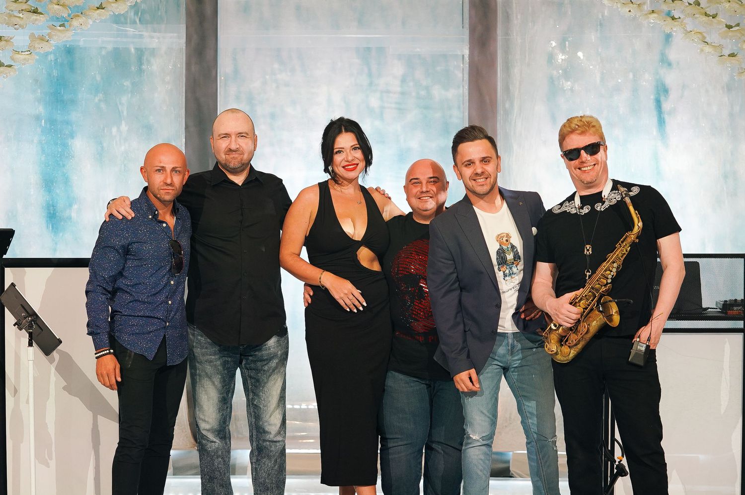 Band members posing together on stage against blue watercolor backdrop at event venue.