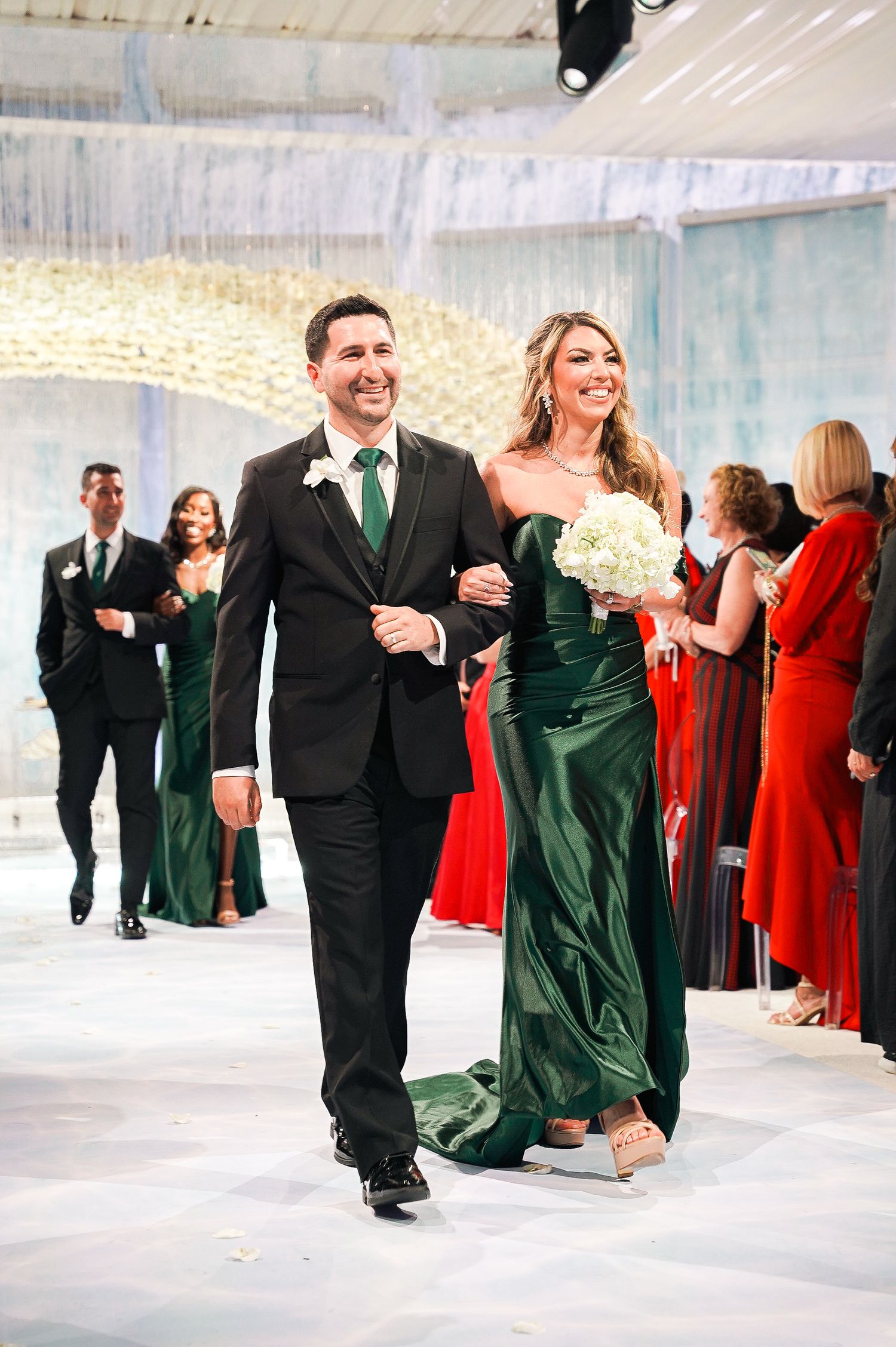 Wedding party members walk down aisle in emerald green and black formal attire.
