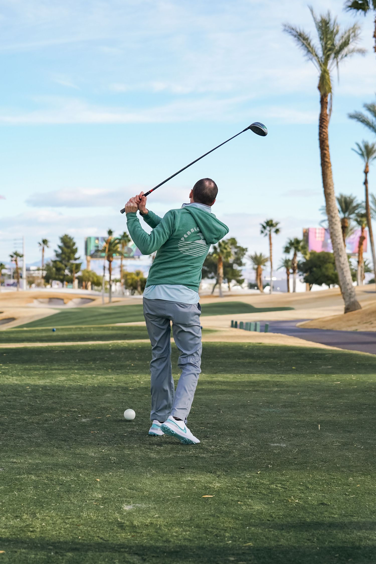 Golfer in green sweater follows through on swing at desert course with palm trees.