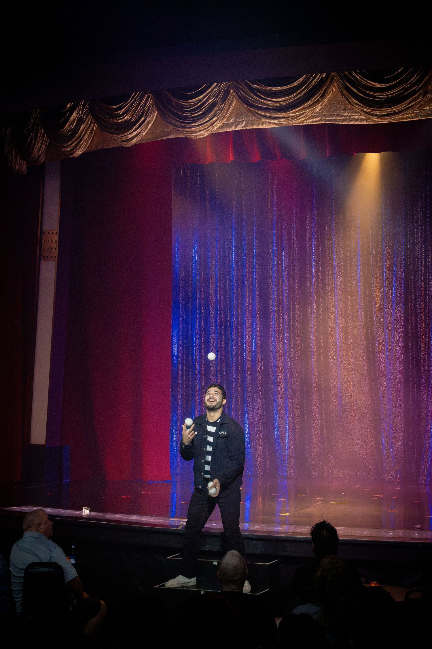 Stage performer with microphone stands against shimmering curtain backdrop under theatrical lighting.