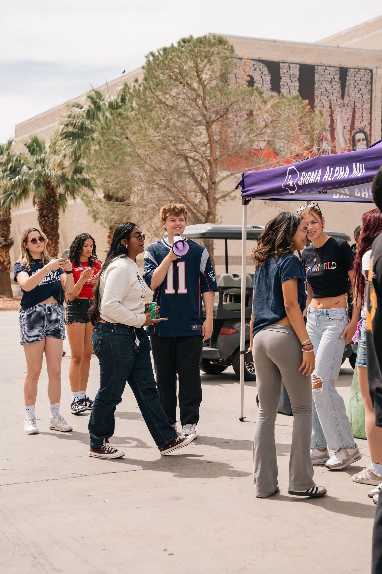Students gathered at an outdoor event with purple tent and campus building in background.