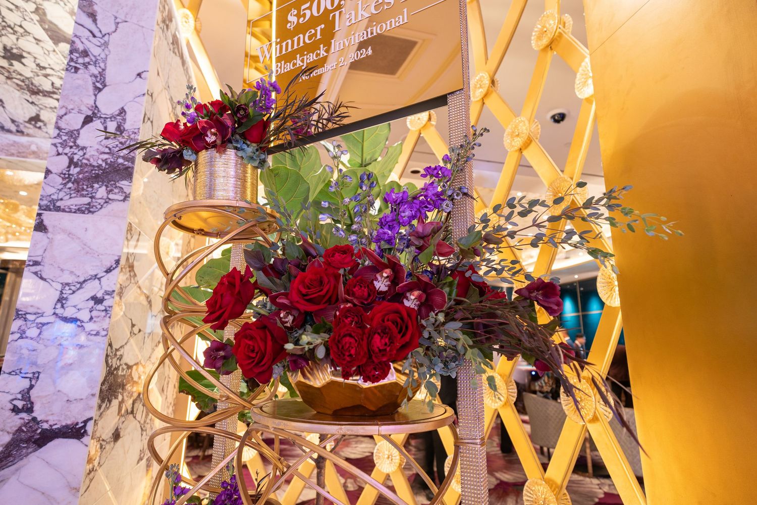 Vibrant floral arrangement with red and purple blooms displayed in an ornate golden vessel against a yellow wall.